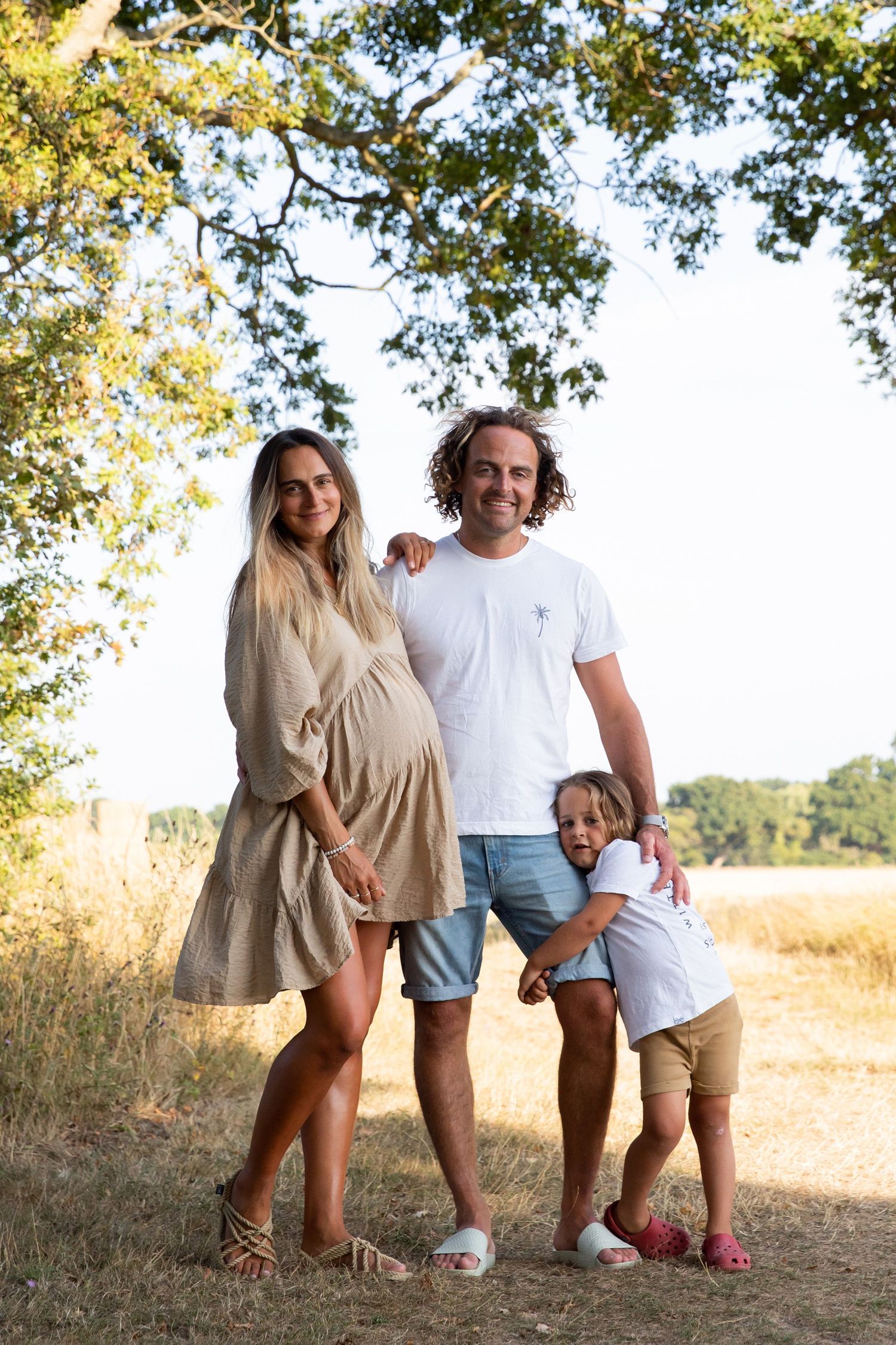 A family portrait outdoors with sun-dappled trees and golden fields in the background.