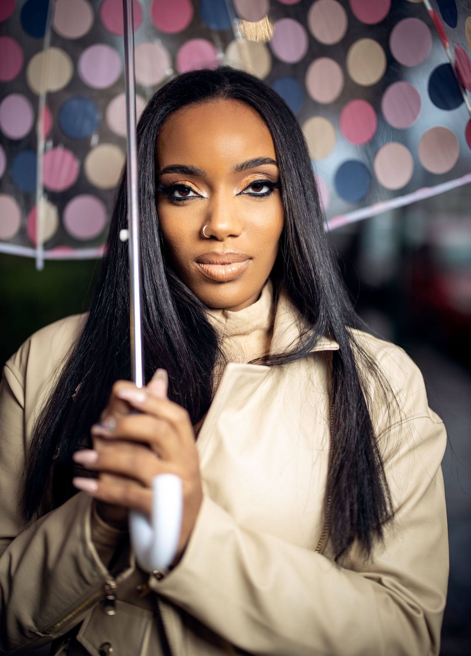 A person with long hair holds a clear polka dot umbrella against blurred lights.