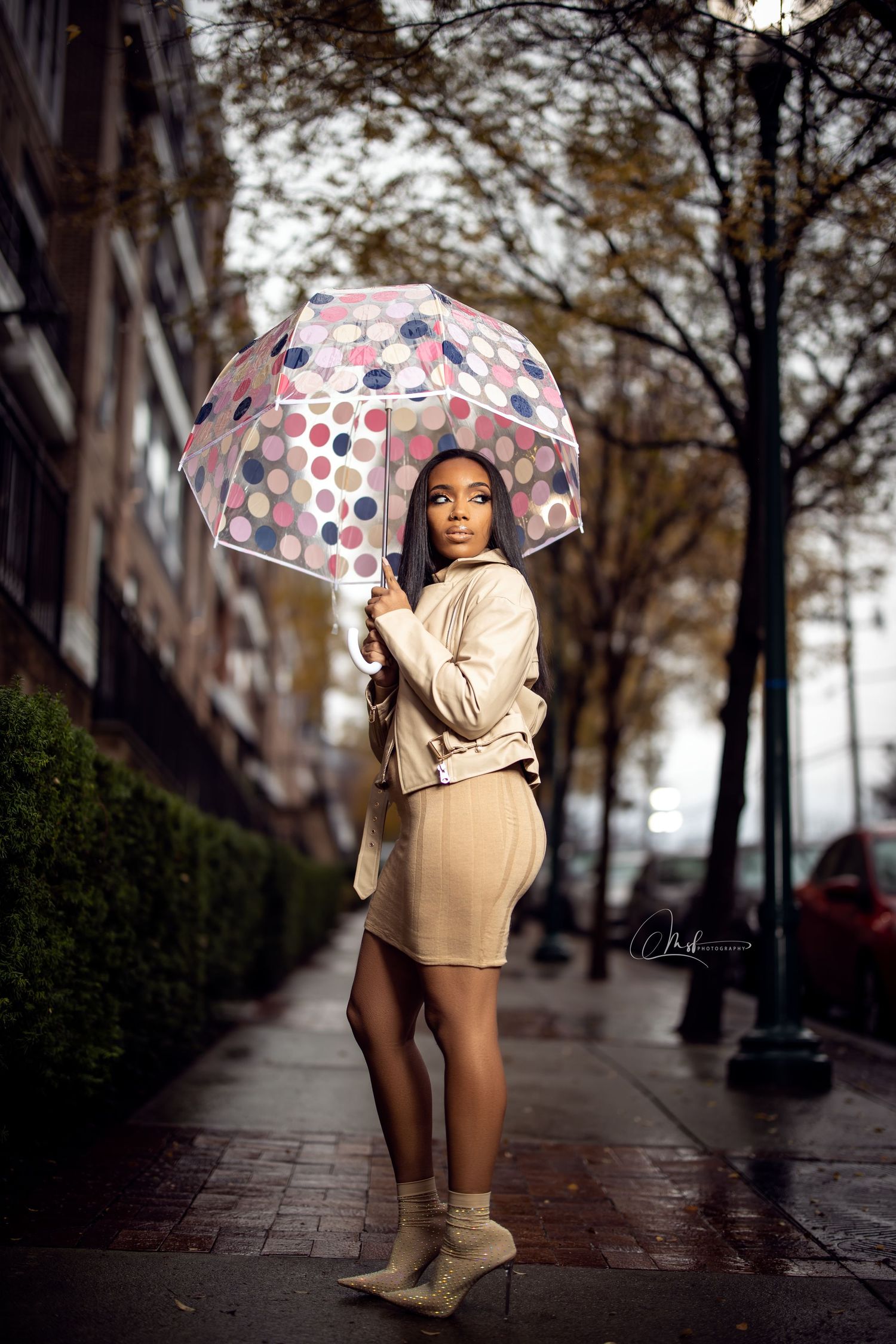 A person in neutral tones poses with a patterned umbrella on a rainy city street.