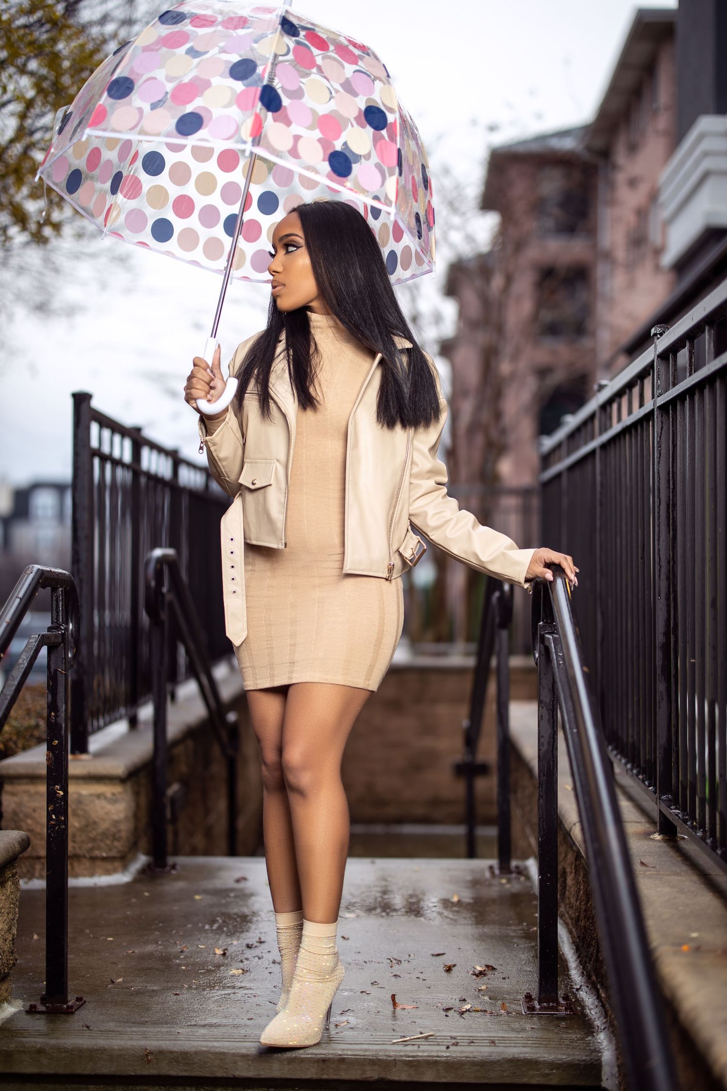 A person in beige outfit holds a polka dot umbrella while posing on wet stairs.