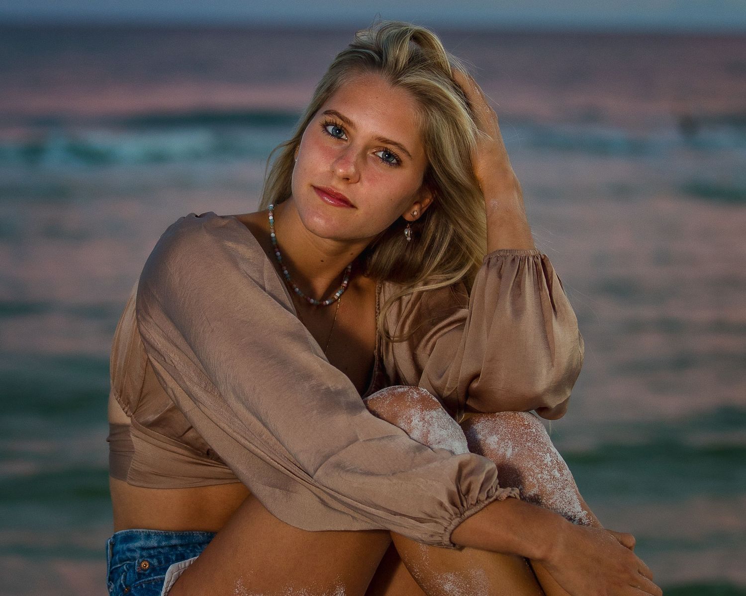 Portrait at sunset showing relaxed pose in tan top and denim against beach backdrop.