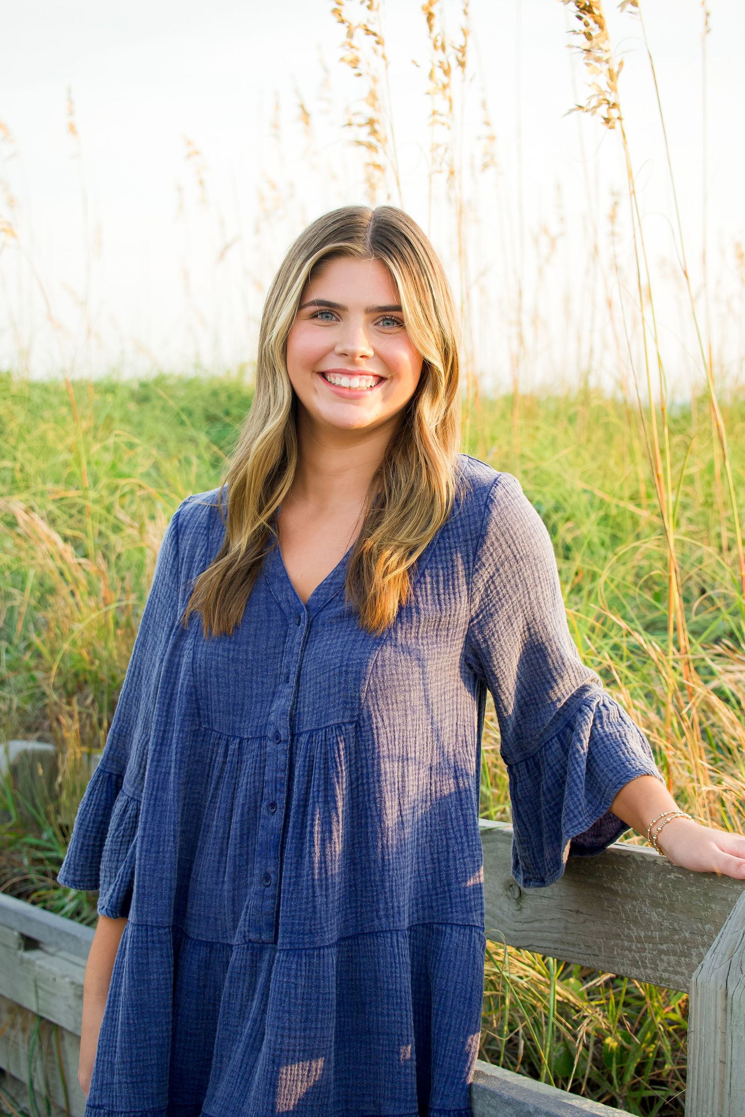 Woman in blue dress stands by wooden fence with tall grass in background.