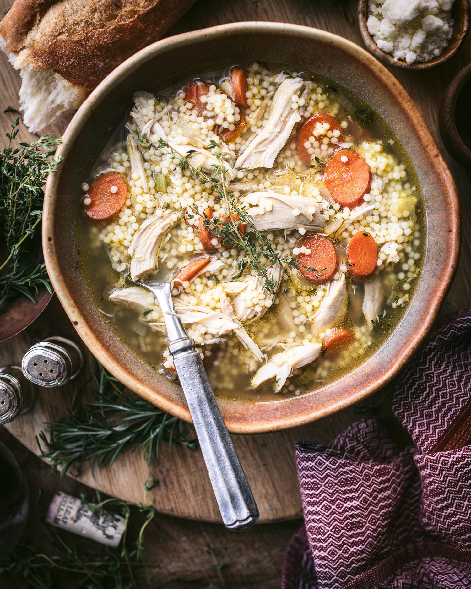 Overhead view of hearty chicken and pearl couscous soup with carrots and herbs in a rustic ceramic bowl.
