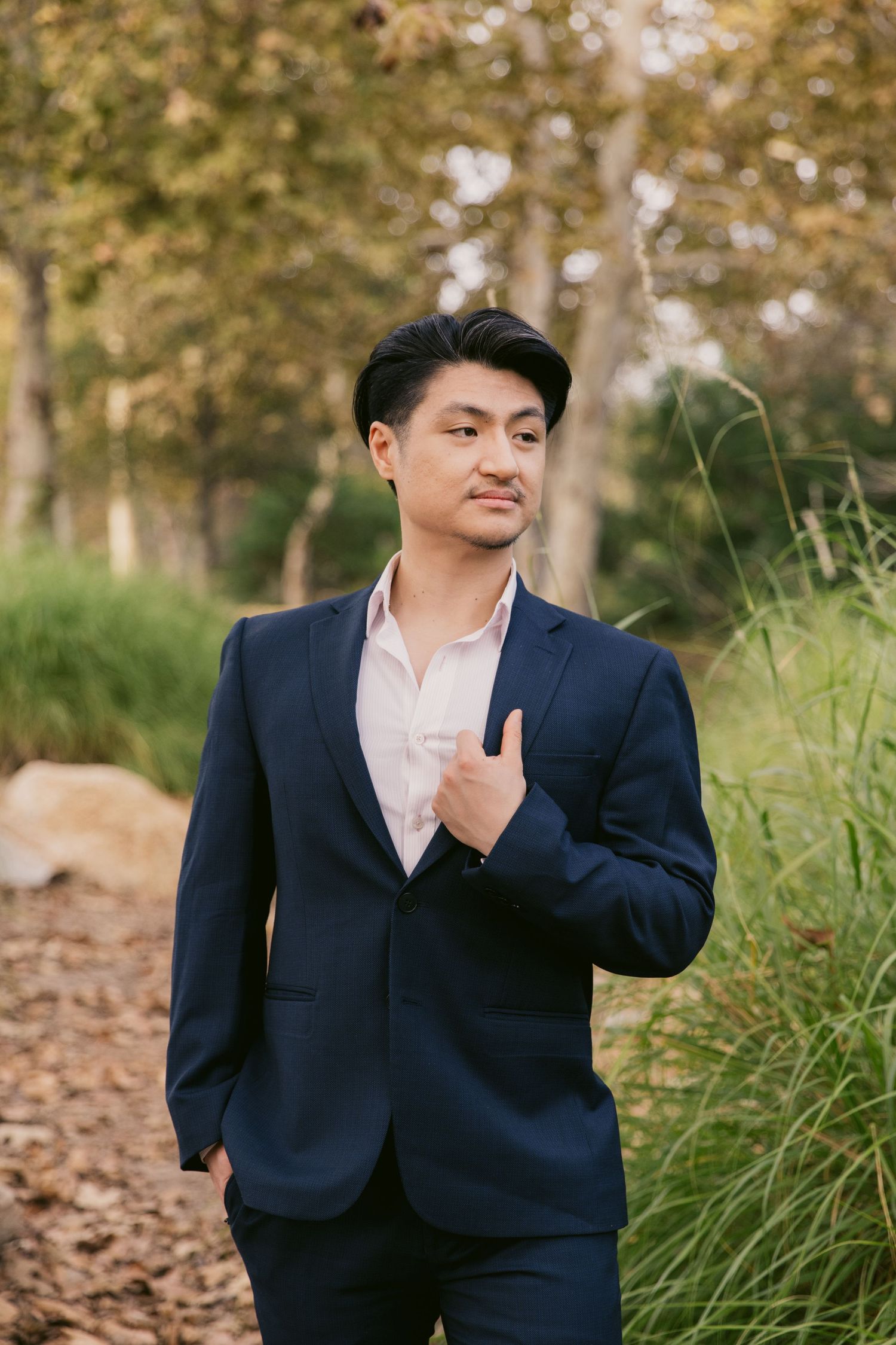 A person in formal attire poses confidently against a backdrop of tall grass and autumn trees.