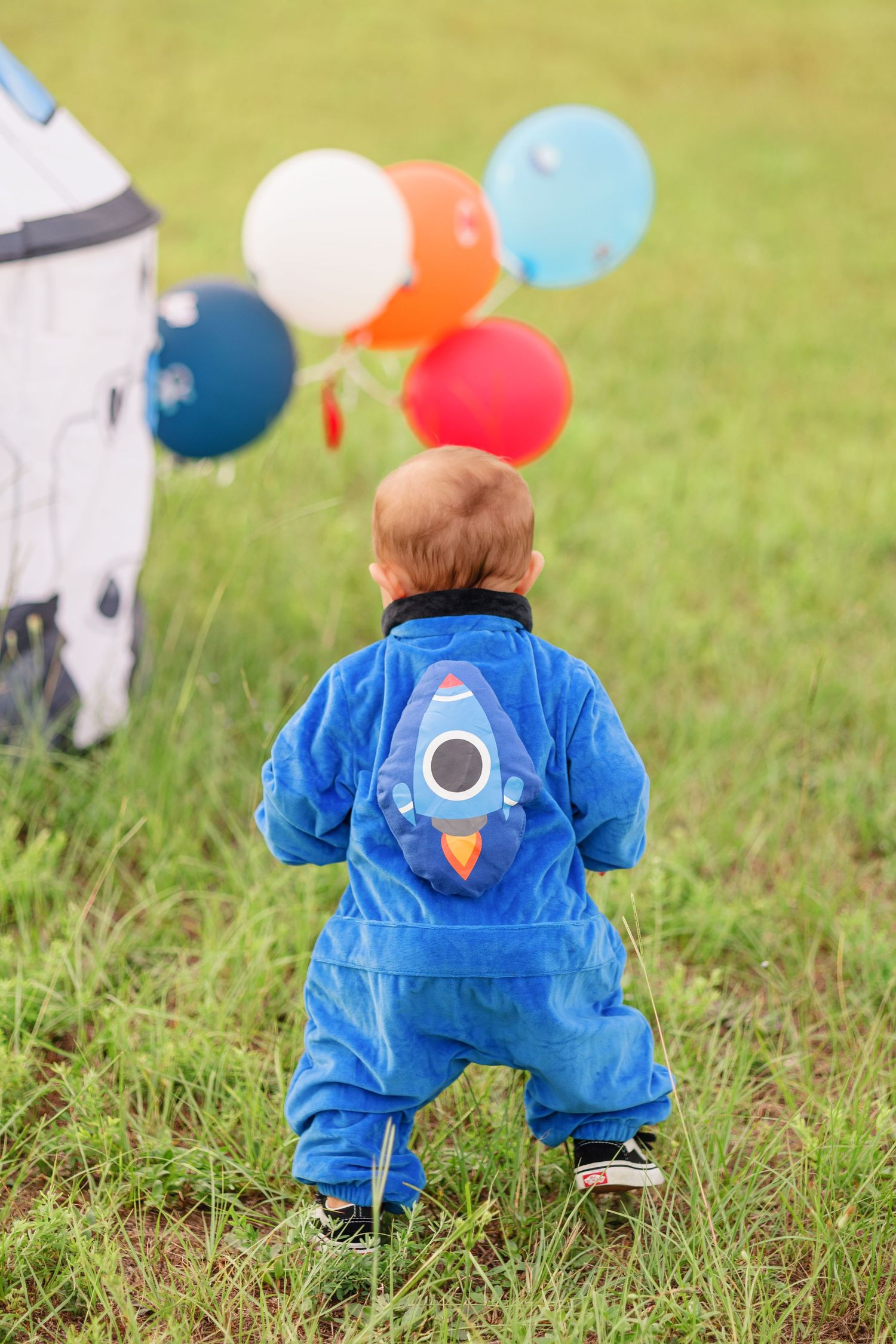 A child in a blue rocket-themed jumpsuit looks at balloons near a space-themed tent.