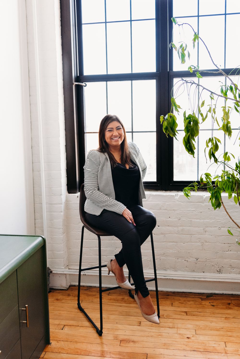 Professional woman in striped blazer sitting on high chair near bright window with plants.