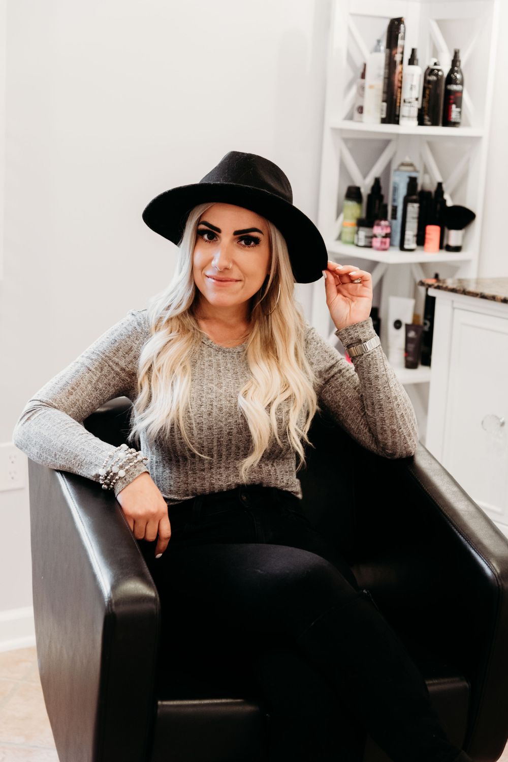 Woman in grey sweater and black hat poses confidently in leather salon chair.