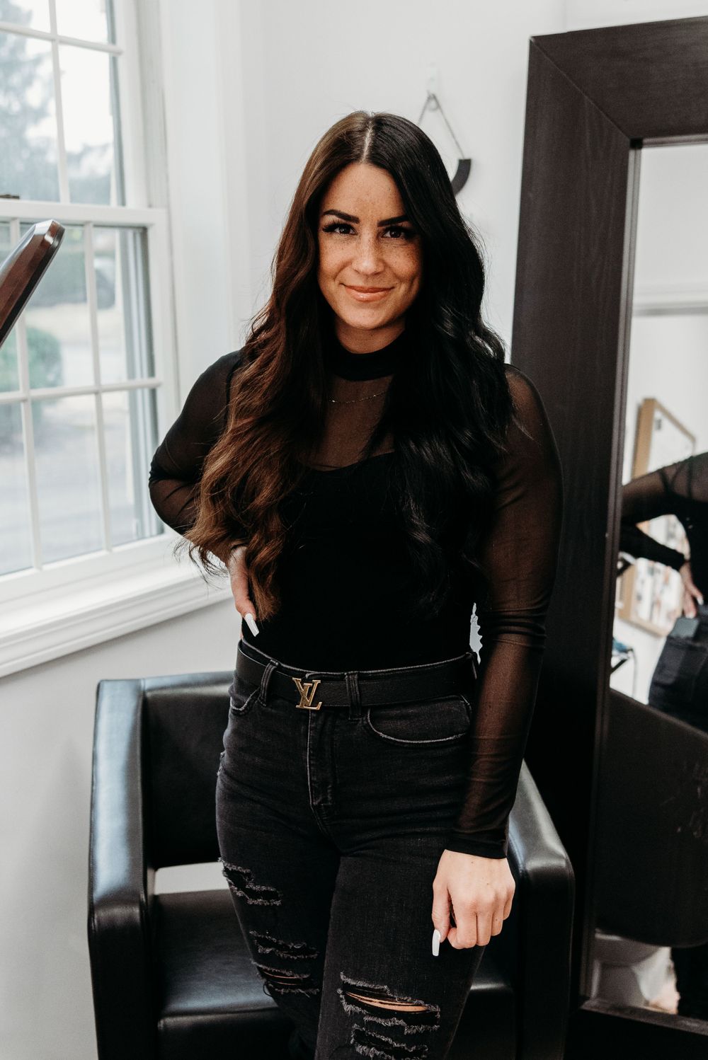 Woman in black mesh top and distressed jeans posing against white wall in salon setting.