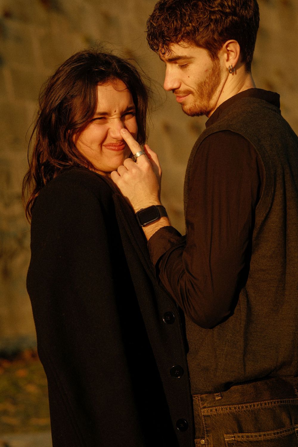 A warm-toned portrait of a couple sharing a lighthearted moment against a stone wall at sunset.