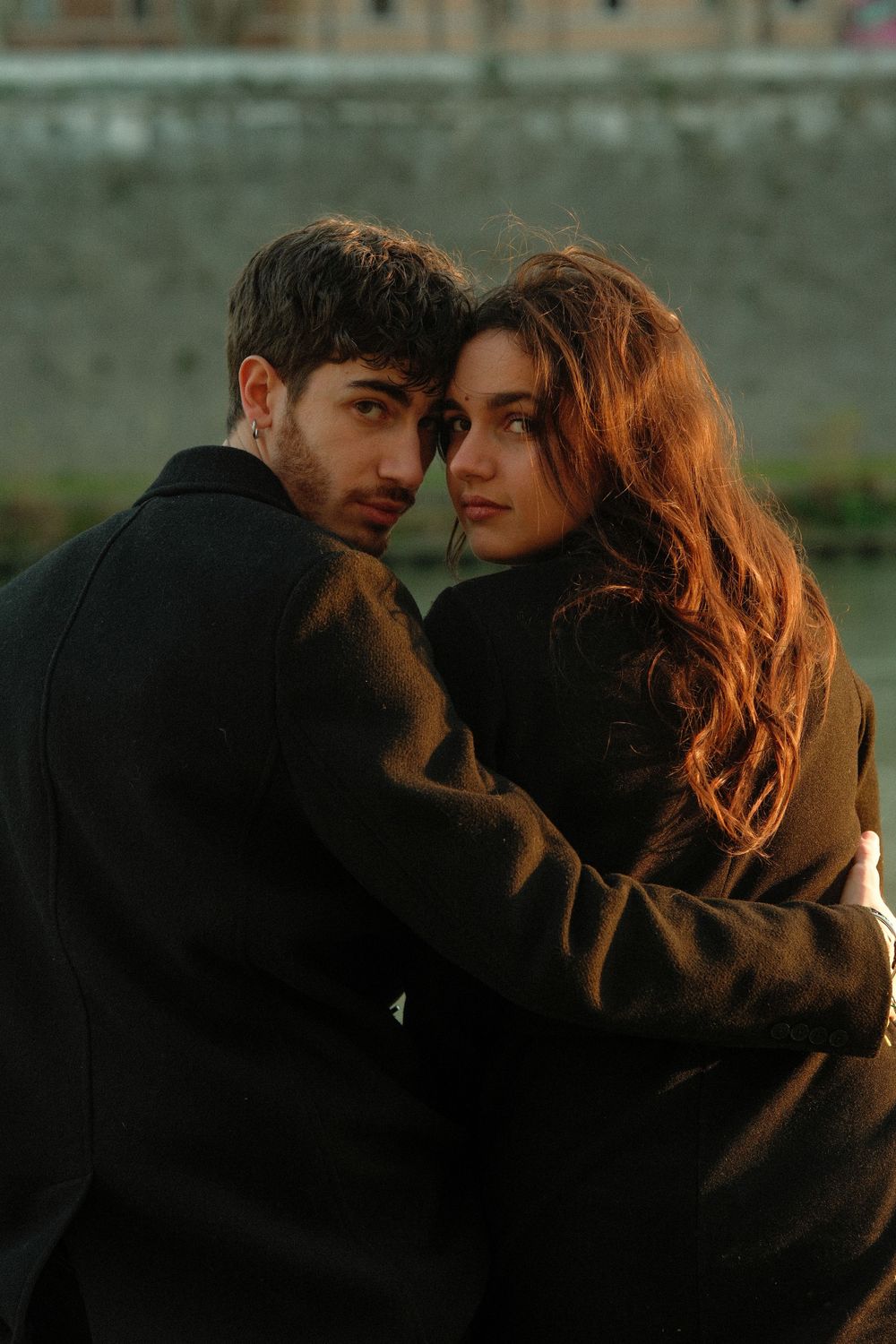 A couple in dark coats shares an intimate moment during golden hour with soft lighting.