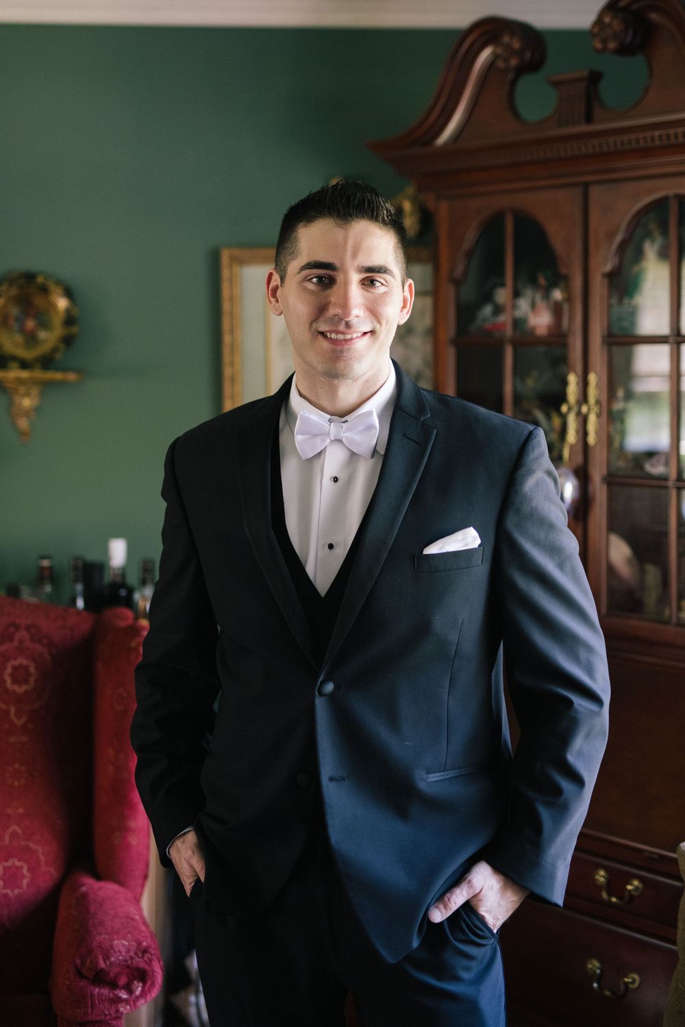 Formal portrait of groom in classic black tuxedo and bowtie standing in elegant room.