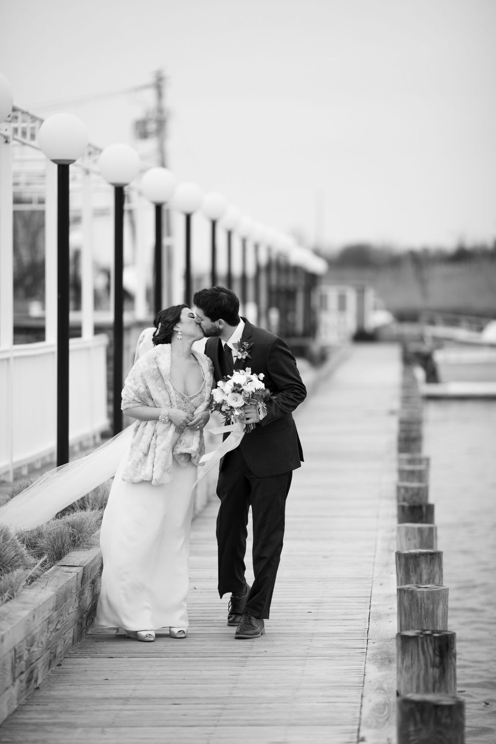 Black and white photo of newlyweds walking intimately along wooden boardwalk.