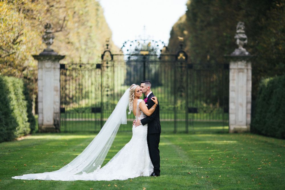 Wedding couple embraces on manicured lawn in front of grand estate gates.
