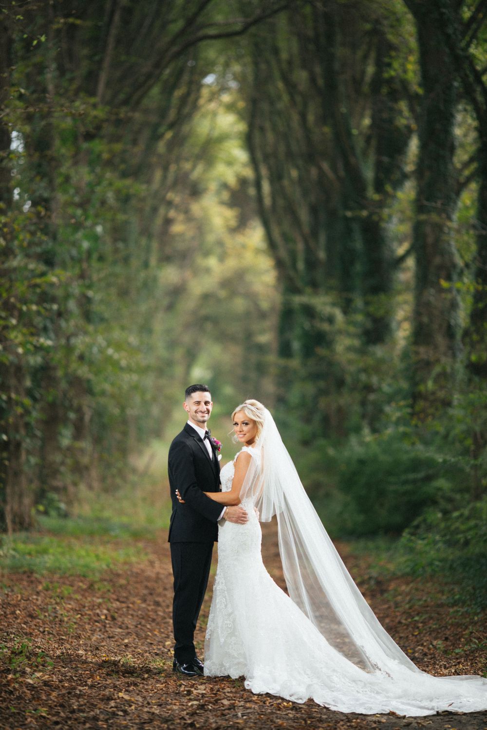 A bride and groom pose for a romantic portrait on a tree-lined path covered in autumn leaves.
