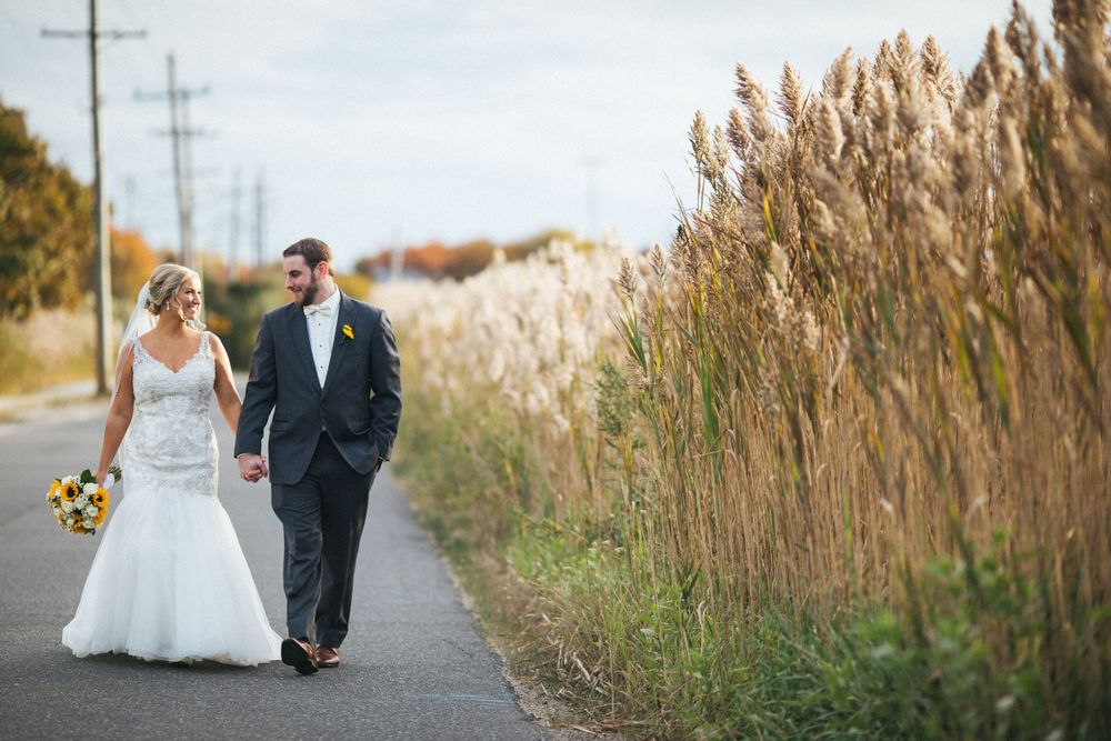 A newlywed couple walks along a country road beside tall ornamental grasses.