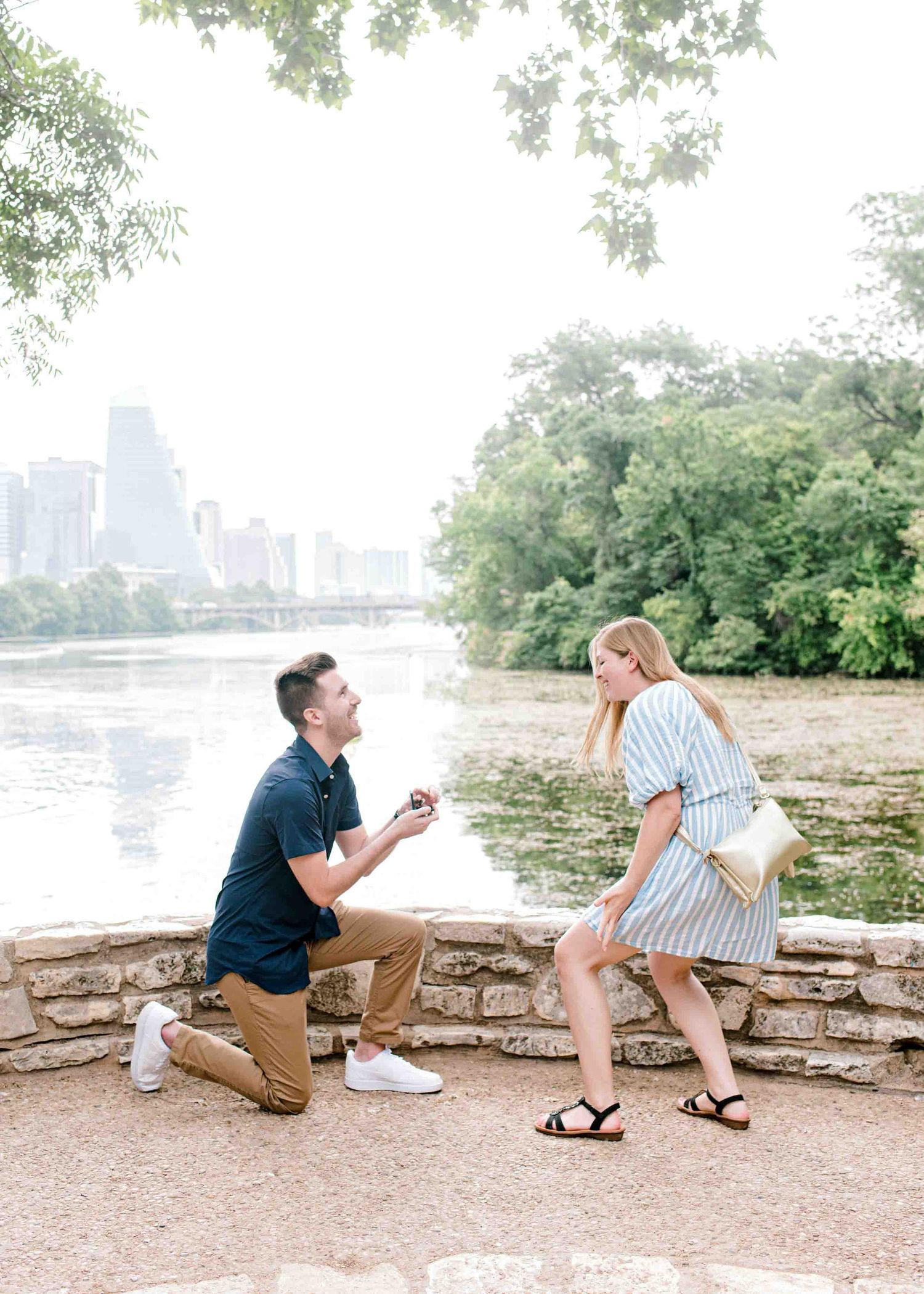 Surprise proposal moment captured beside scenic river with downtown skyline.
