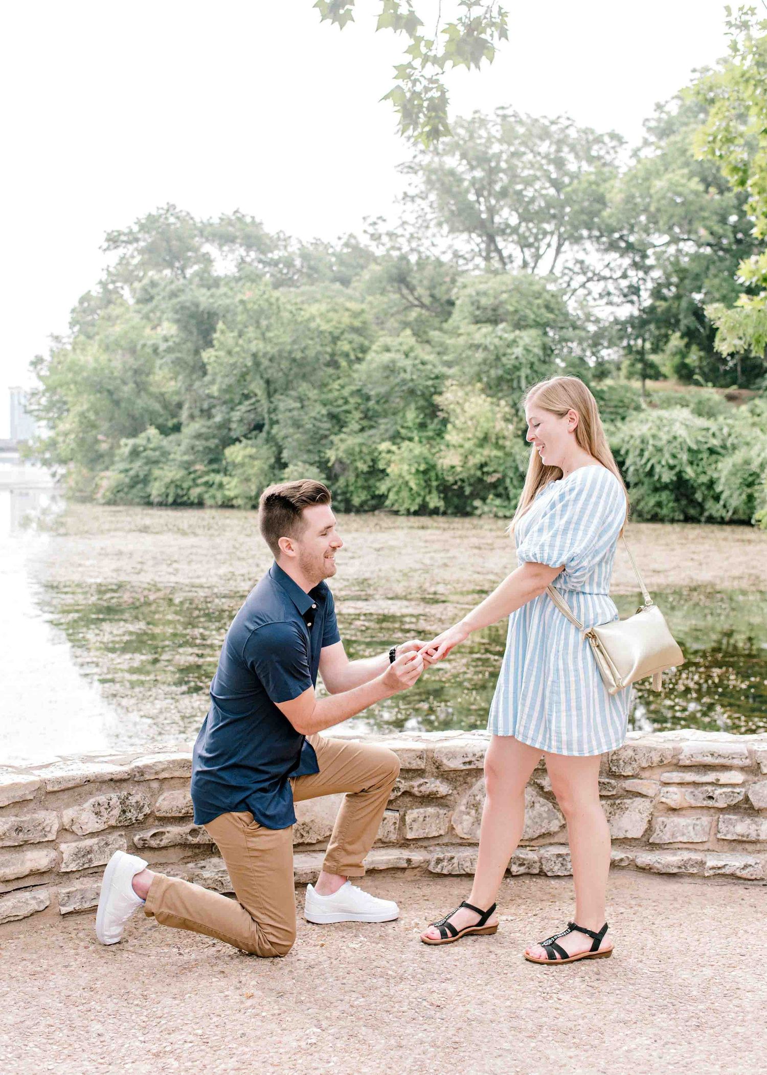 Man kneeling to propose on stone path with river and trees in background.