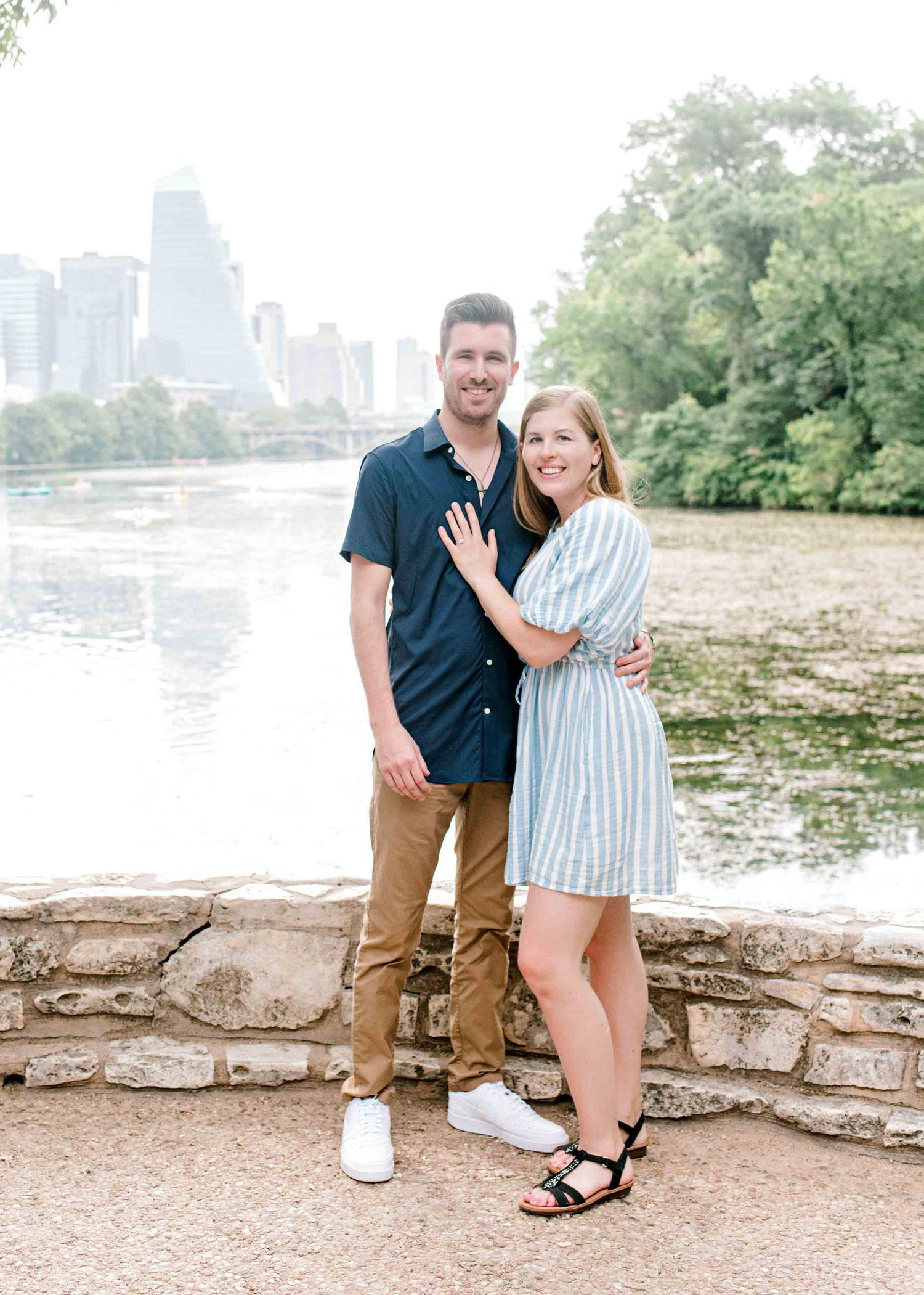 A couple poses together on a stone wall with a city skyline and river in the background.