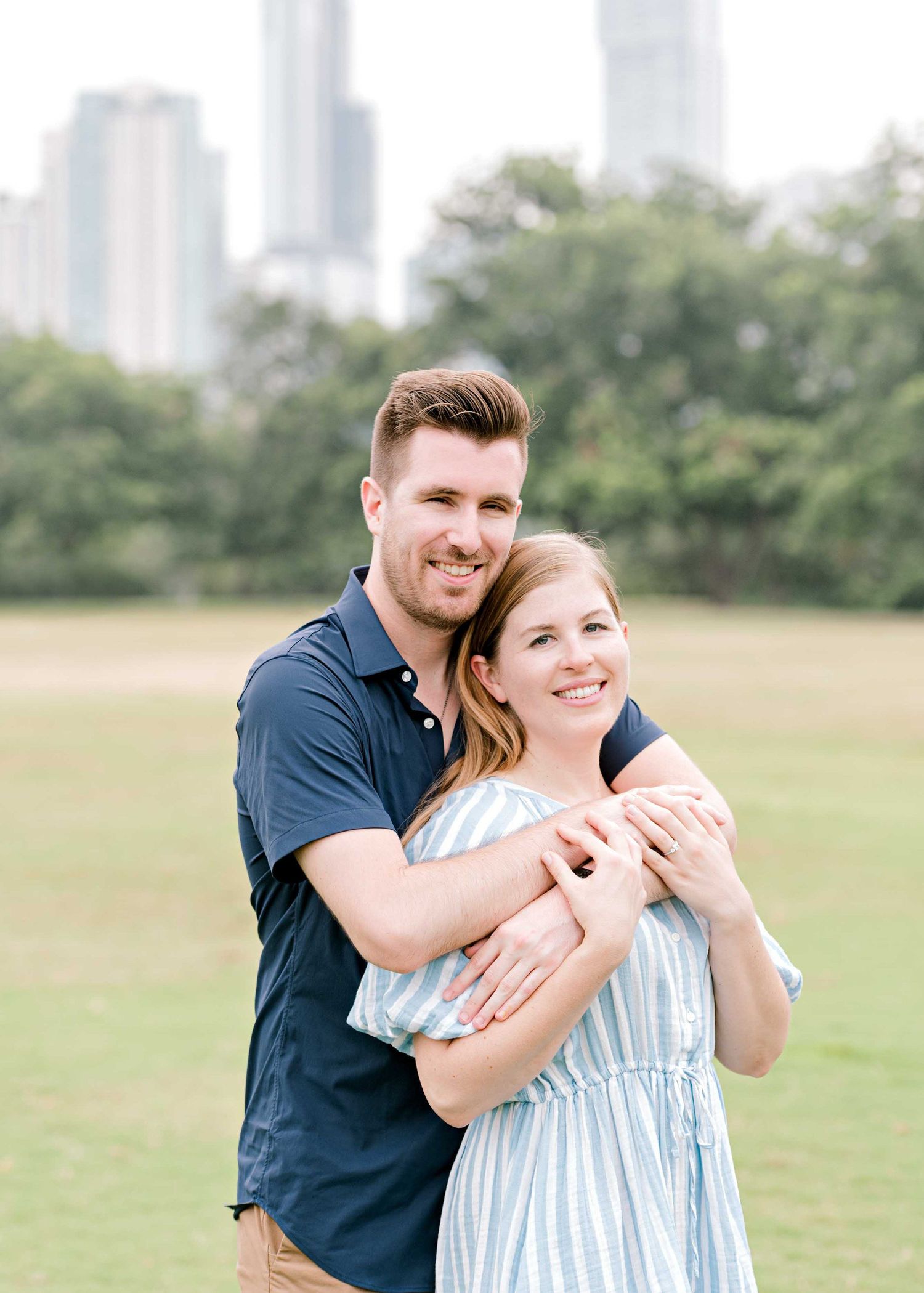 A couple embraces in a park with tall city buildings visible in the background.