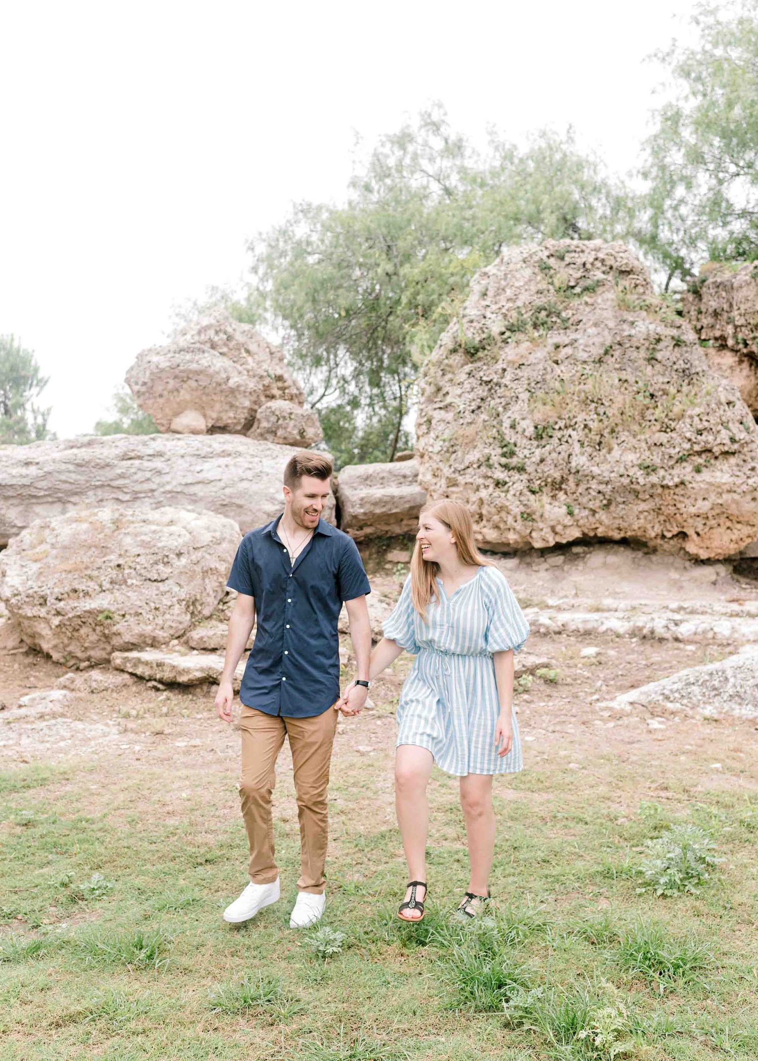 A couple stands together in casual outfits against a rocky natural backdrop on a grassy hill.