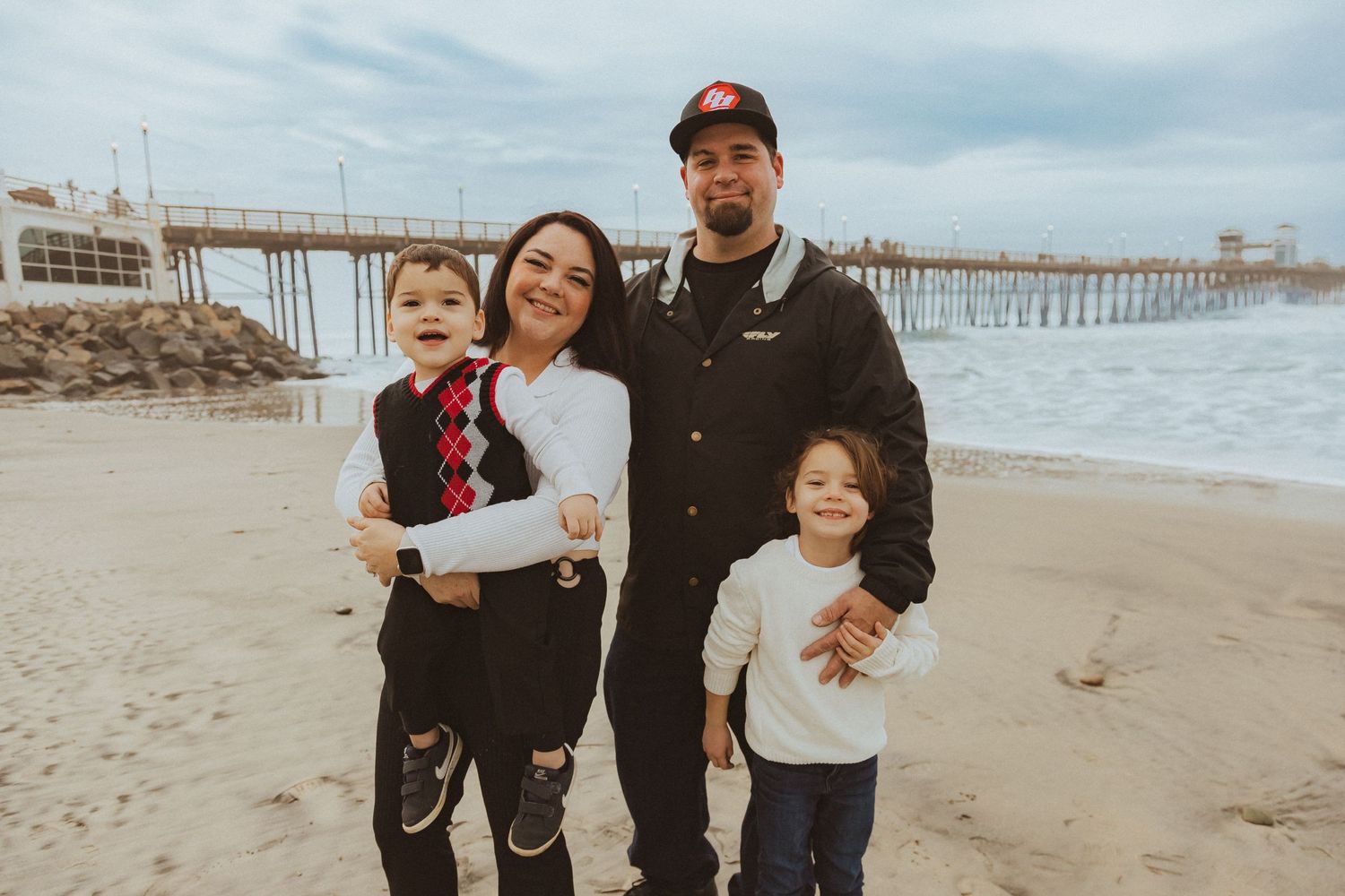 Family posing together on beach with pier visible in background.
