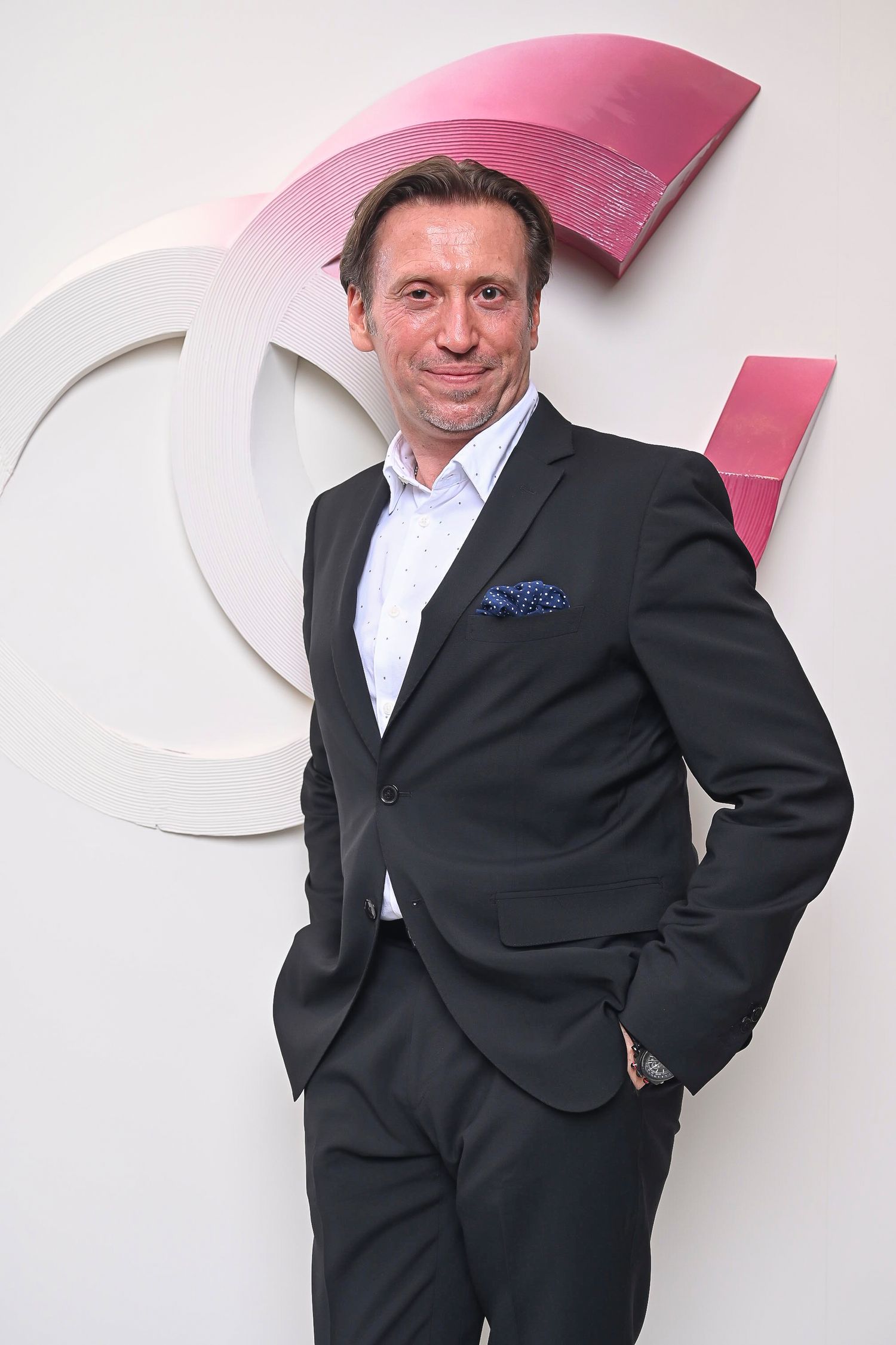 Man in dark suit and blue pocket square poses against pink and white branded wall.