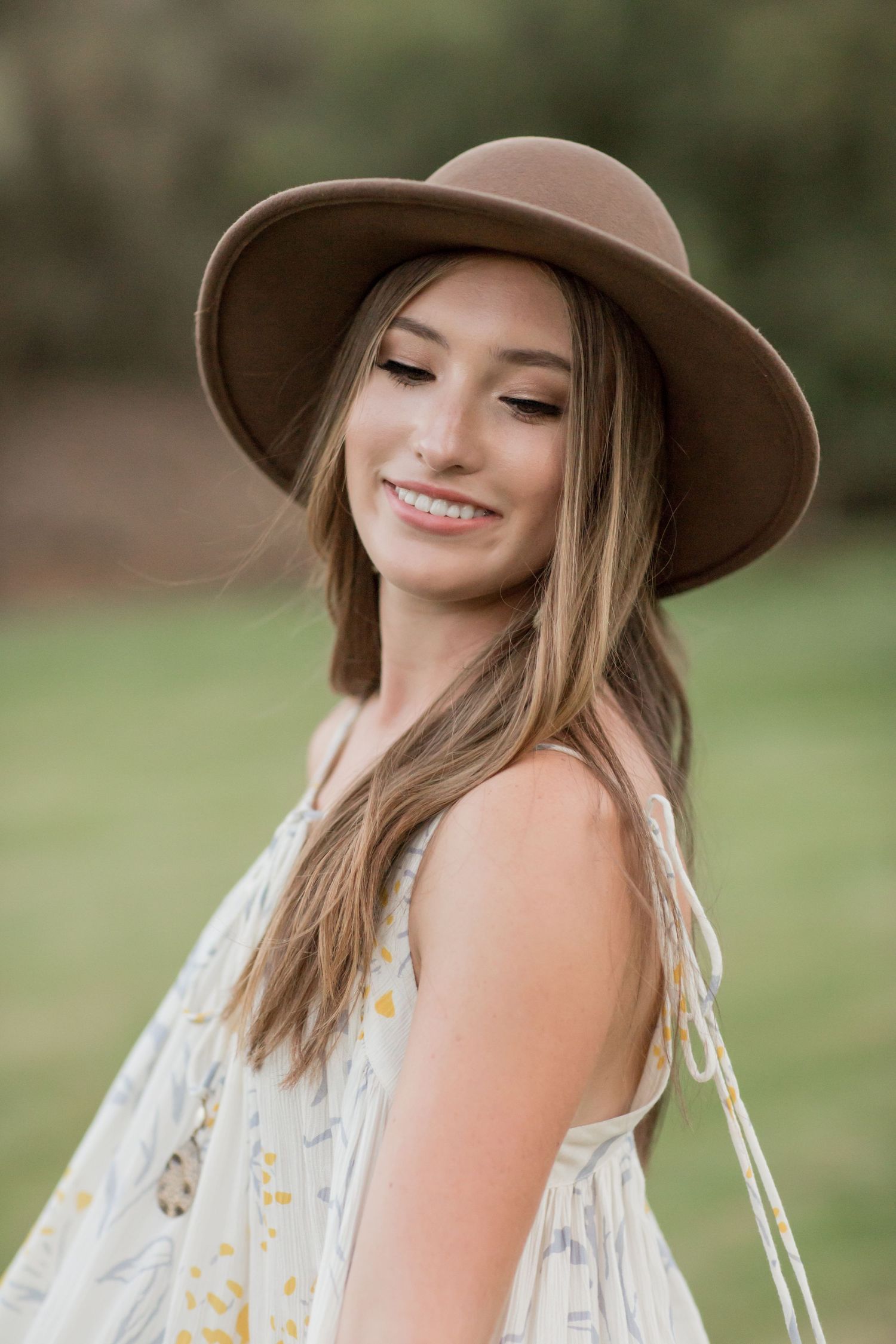 Smiling young woman in floral dress and brown wide-brim hat posing outdoors in a green field.
