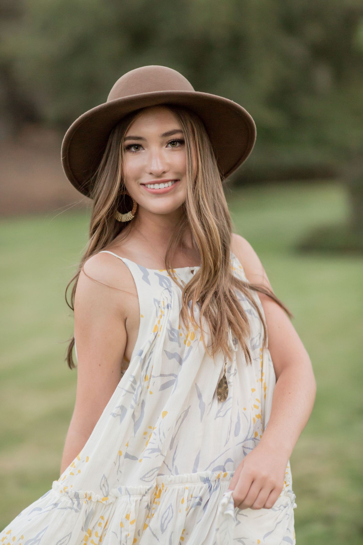 Beautiful young woman wearing a brown hat and floral sundress smiling in an outdoor park setting.