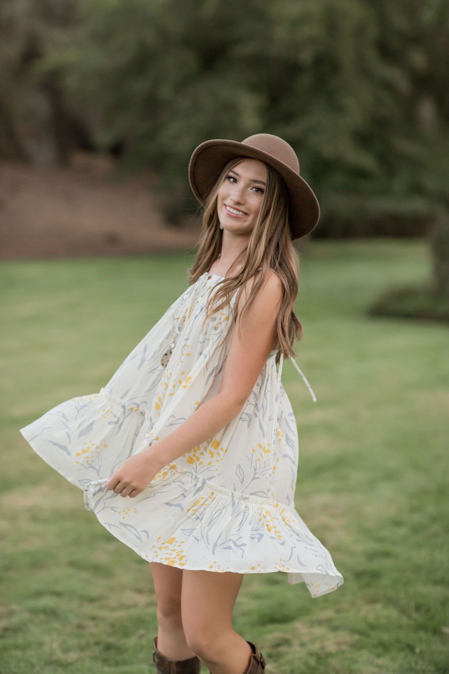 Young woman in white floral dress and brown hat twirling outdoors on a lush green lawn.