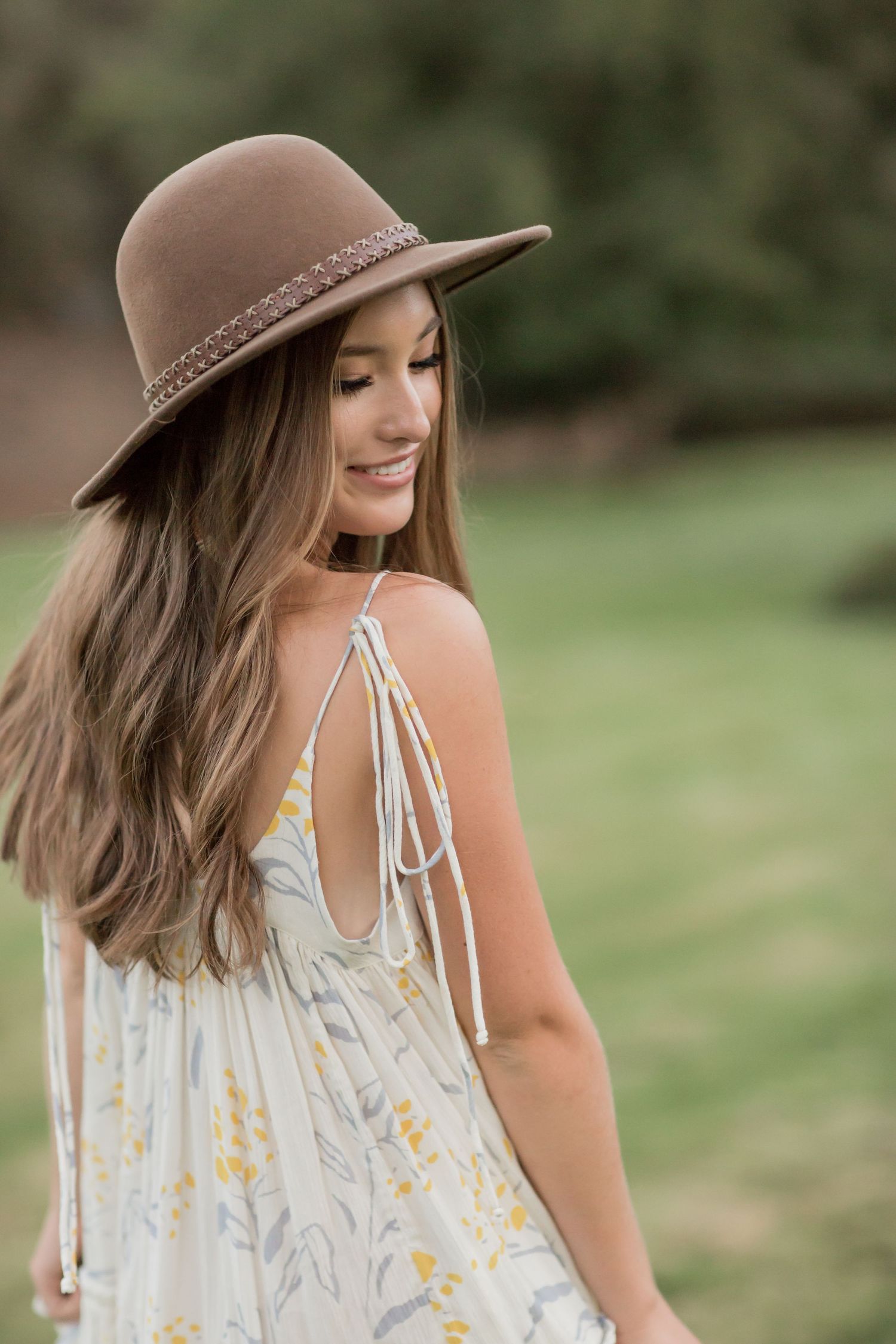 Young woman in white floral sundress and brown braided hat looking down over her shoulder outdoors.