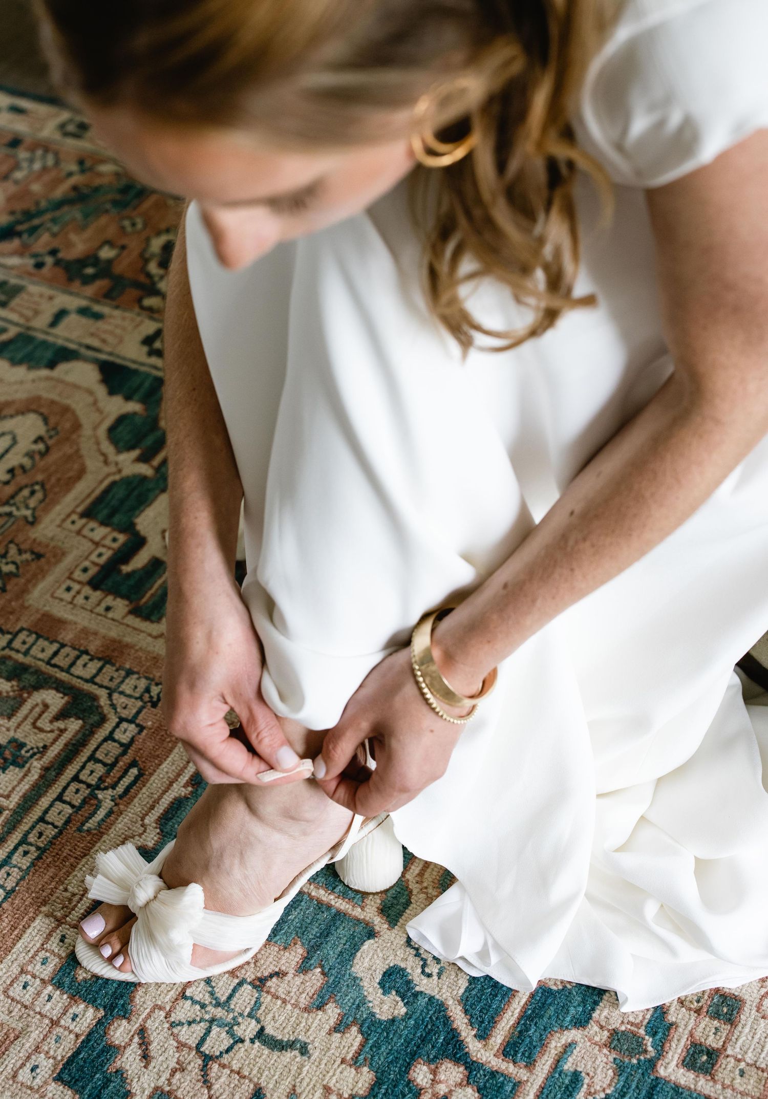Close-up of someone in a white dress adjusting their wedding shoes on a patterned rug.
