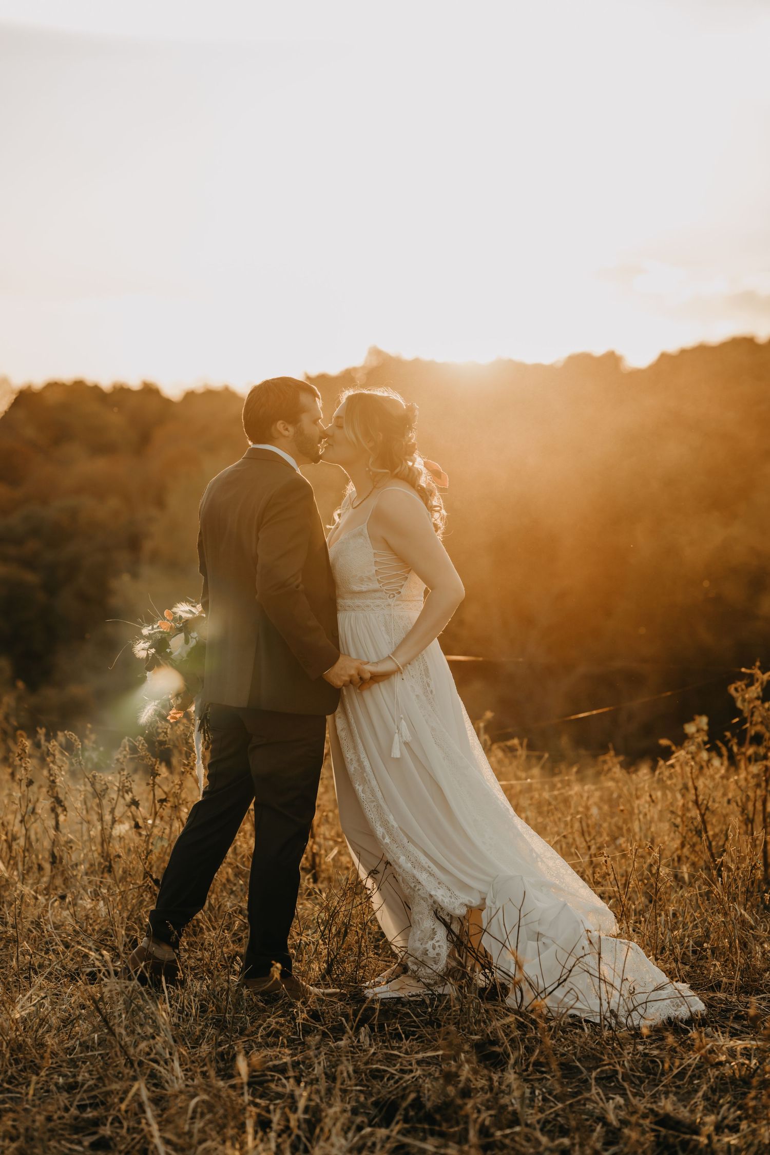 Bride and groom kissing in golden sunset light in a meadow, romantic outdoor wedding portrait.