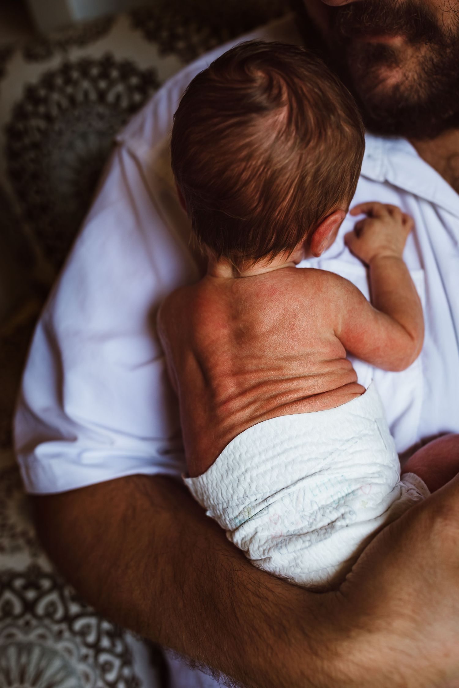 Close-up of a baby in white diaper being held against someone wearing a white shirt.