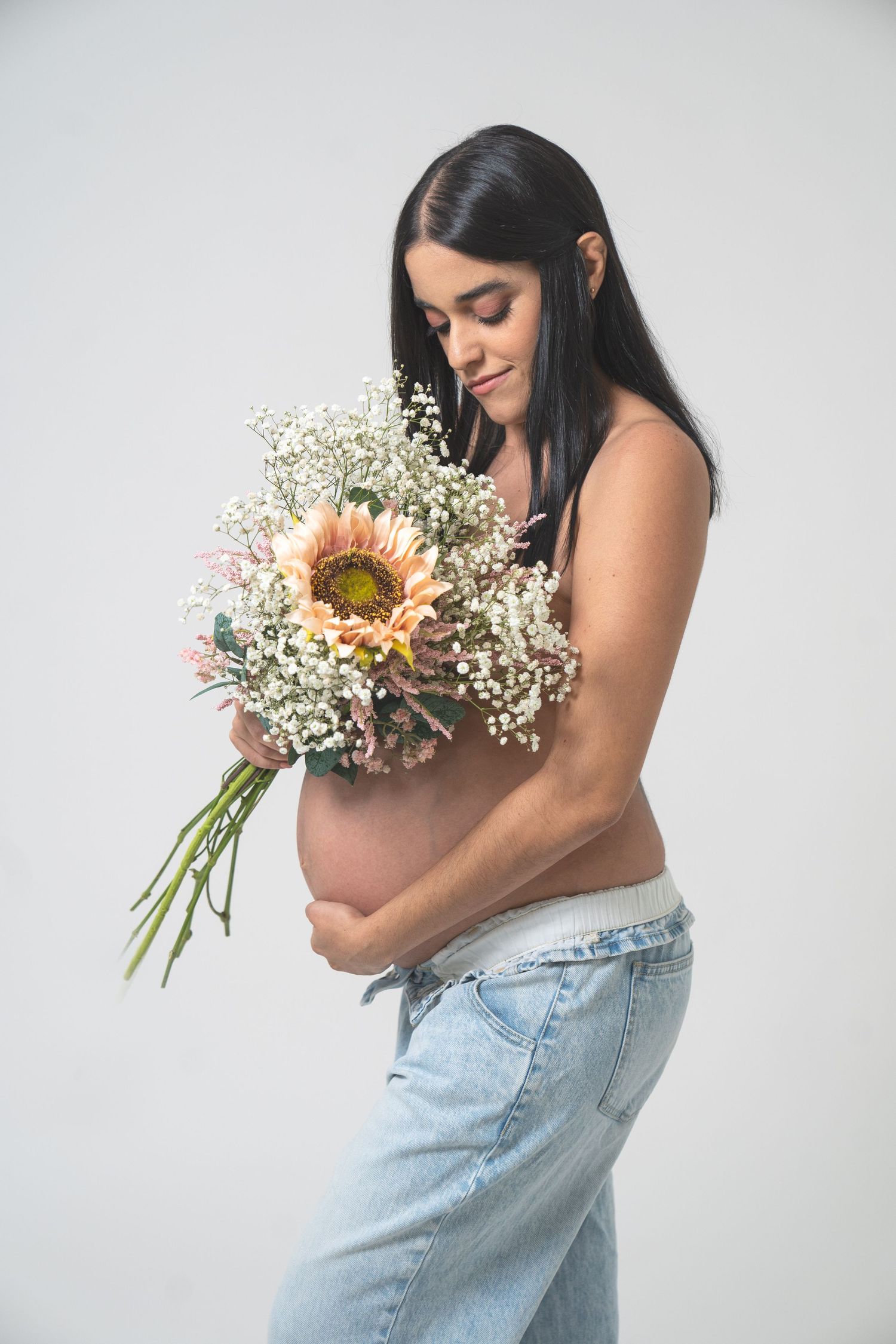 Expectant mother in jeans holds a bouquet of sunflowers and baby's breath against white background.