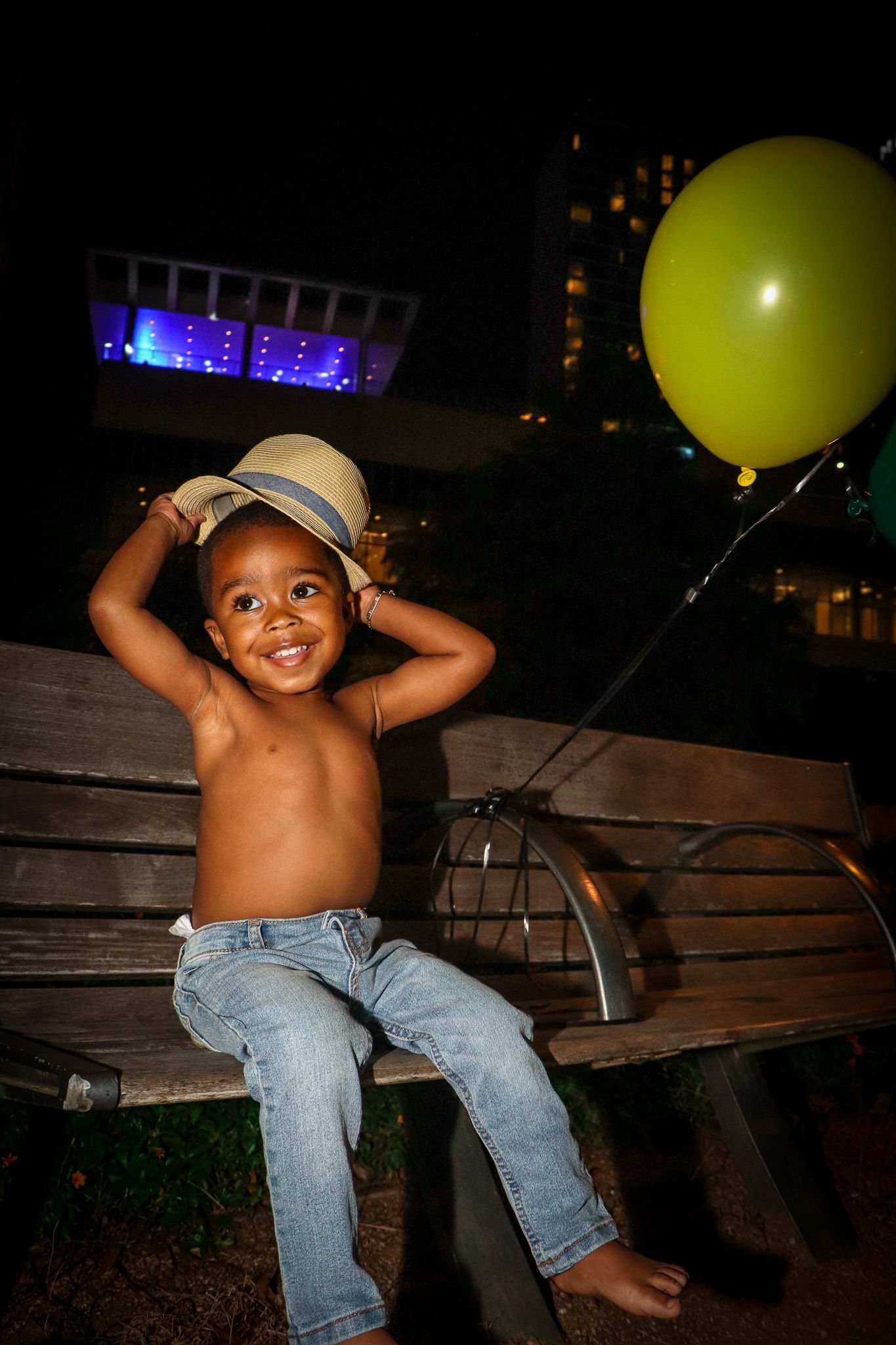 Young child in jeans and hat sits on bench with yellow balloon at night.