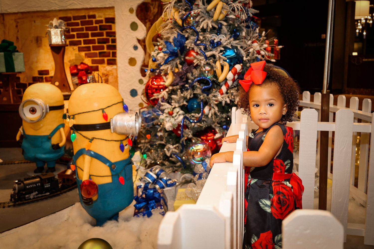 Young child poses near decorated Christmas tree with Minion characters in holiday display.