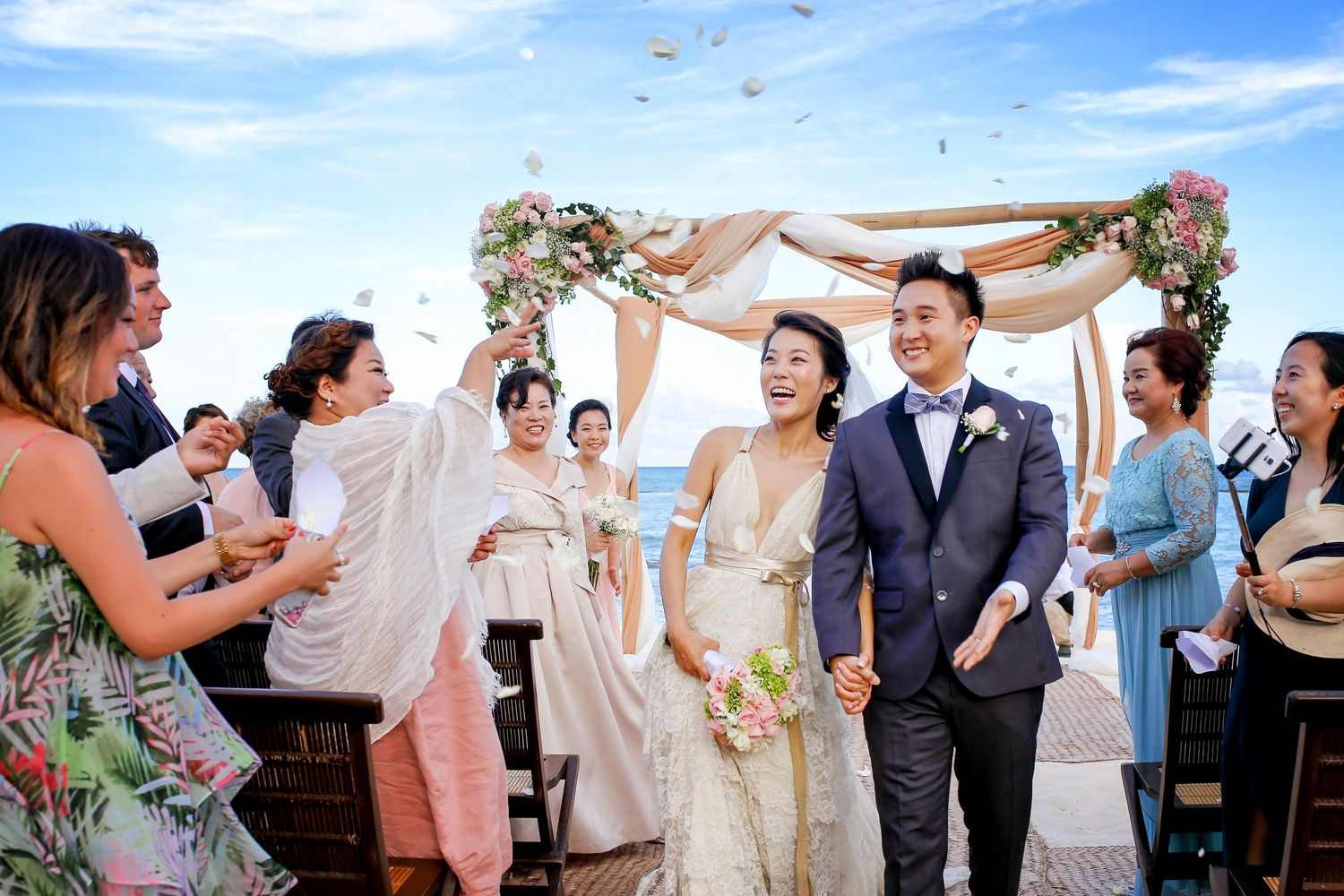 Wedding celebration on a beach with floral arch and guests throwing flower petals during a joyful ceremony.