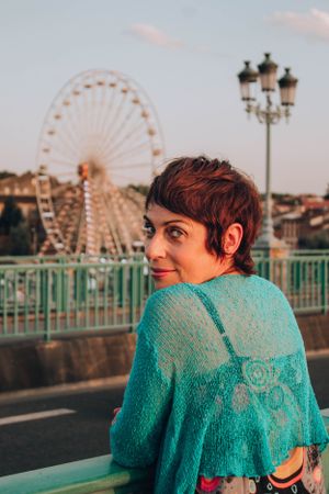 Woman in turquoise sweater overlooking ferris wheel at sunset.