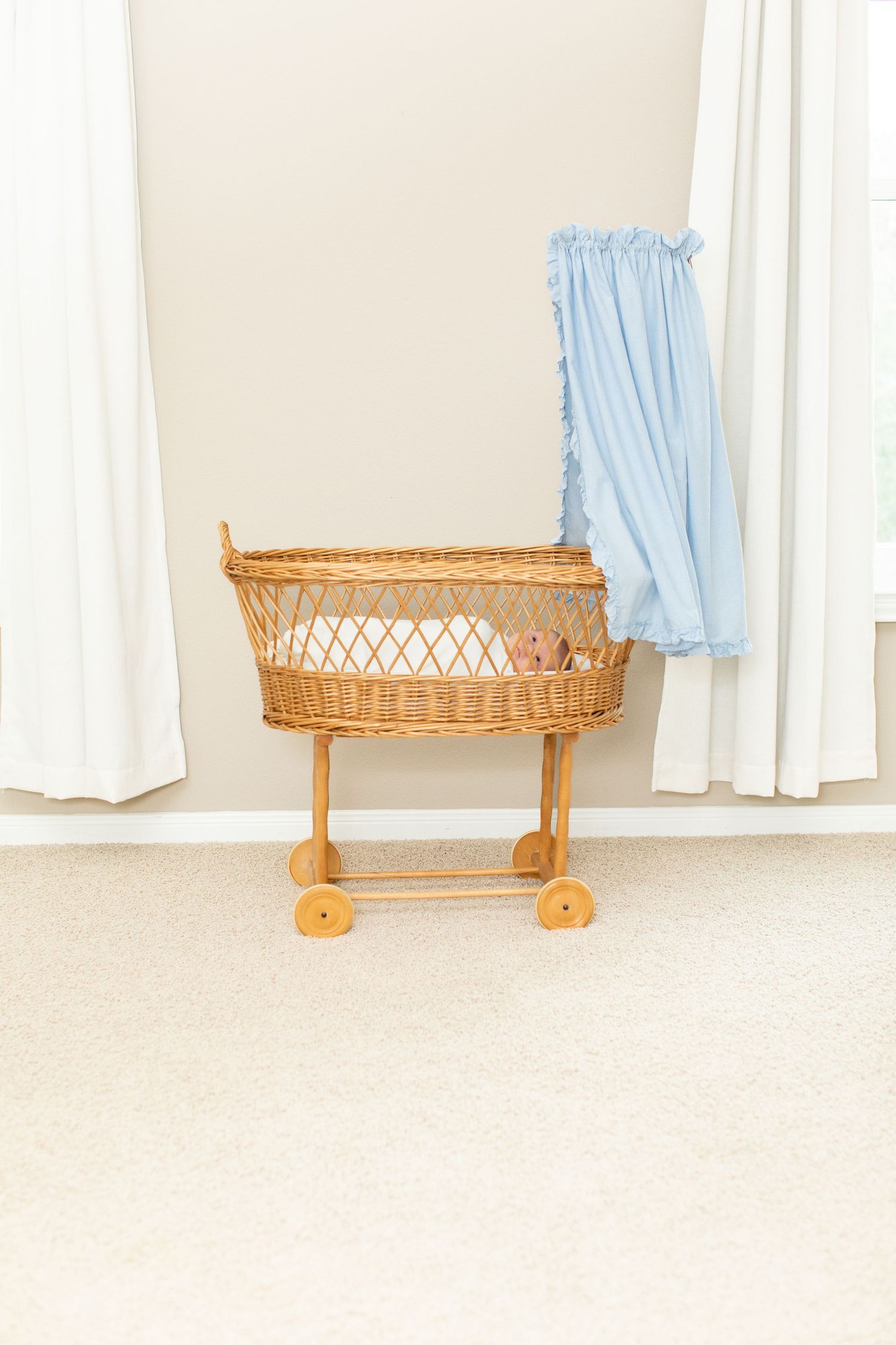 Vintage wicker bassinet with blue fabric and wooden wheels in front of white curtains.