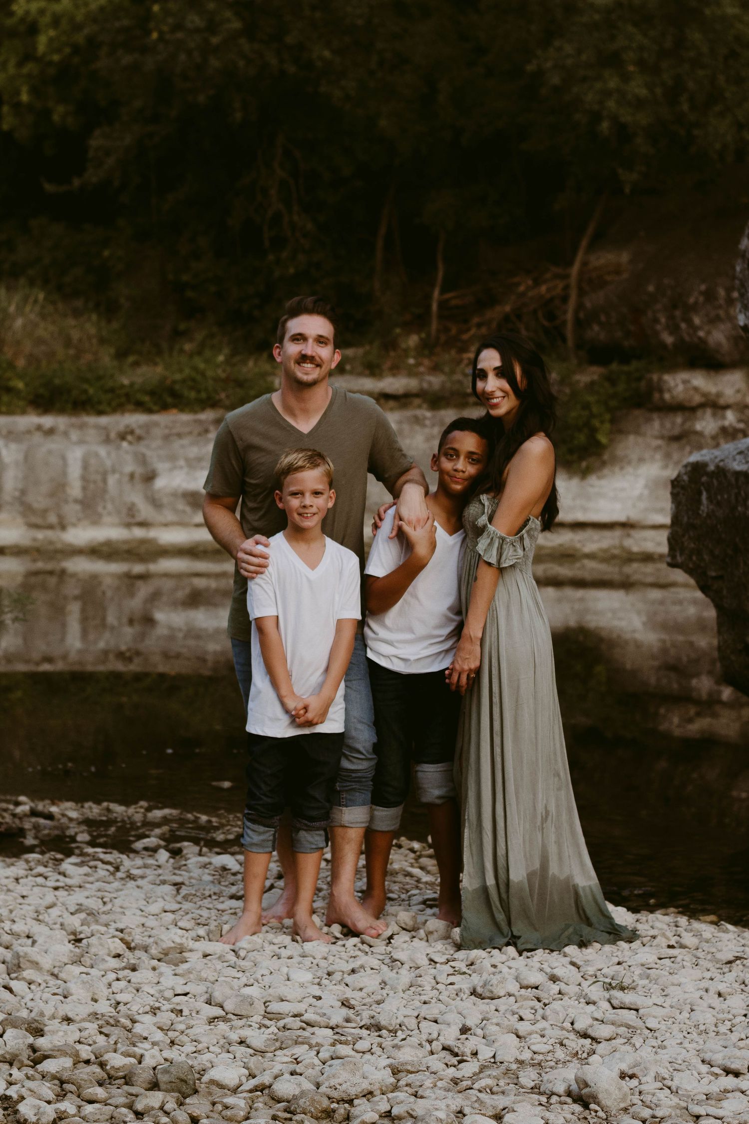 Family of four standing together barefoot on rocky riverbank in casual summer attire.