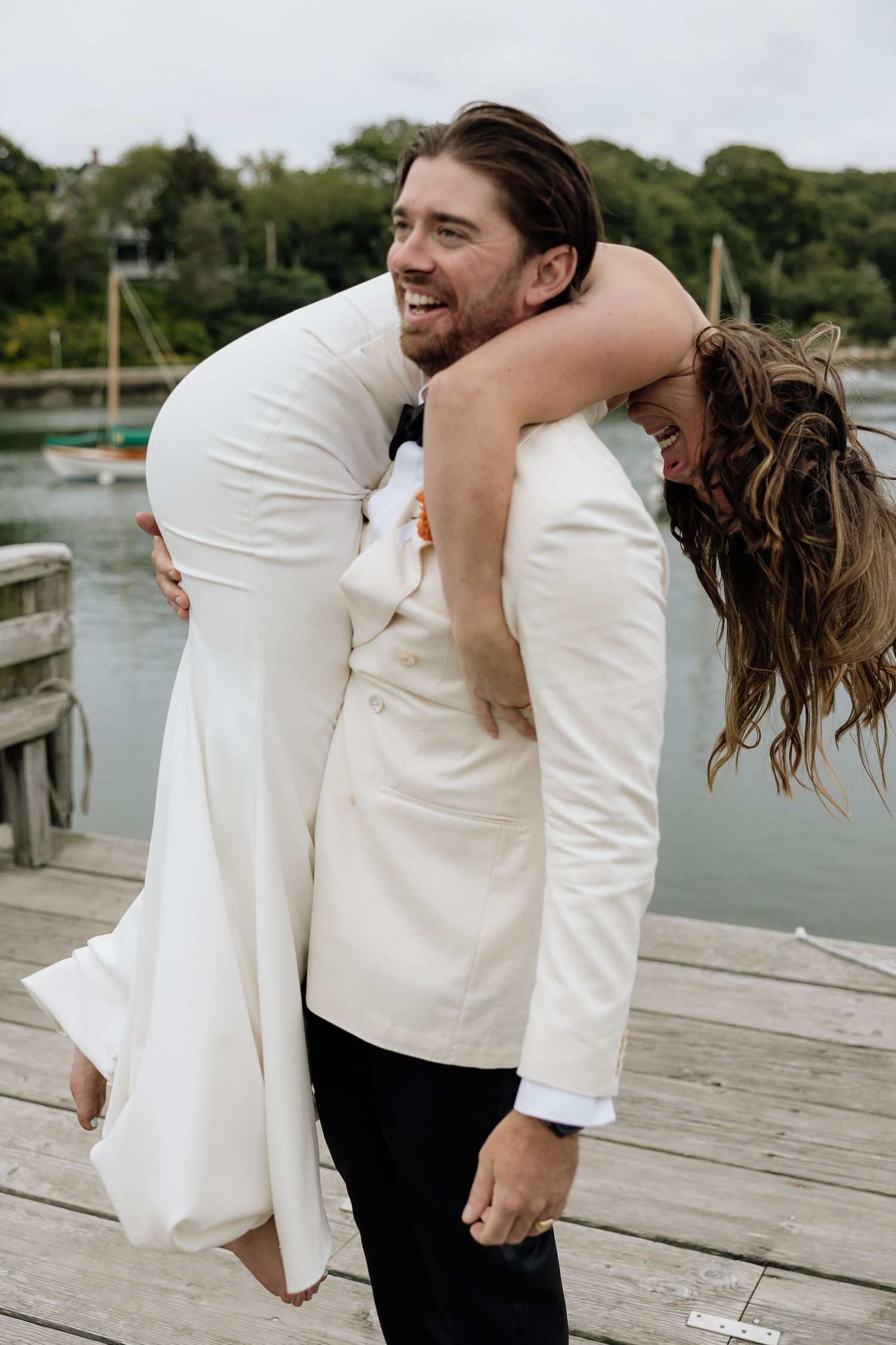 Laughing groom in white suit giving bride a piggyback ride on wooden dock near water. Falmouth, Ma. Miranda Photo & Co