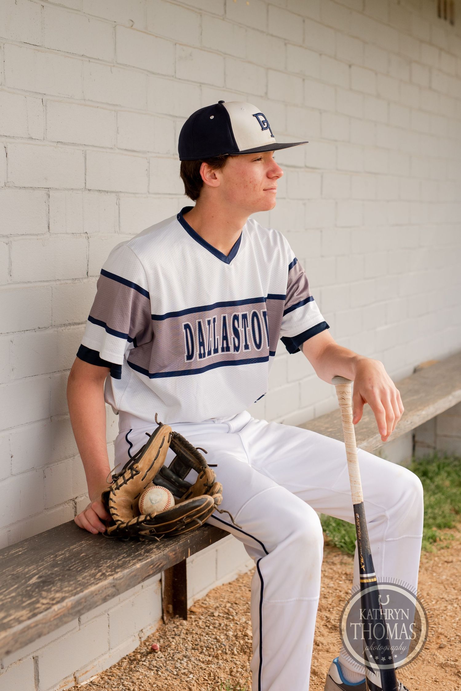 Baseball player in Dellasoft uniform sits on dugout bench with glove and bat.