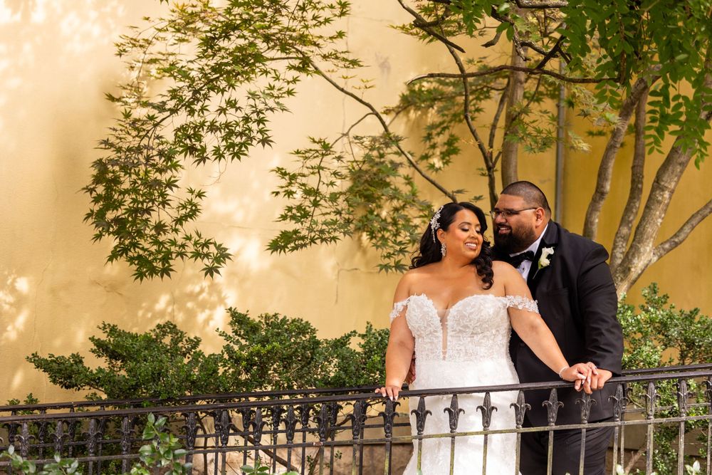 Elegant couple poses on ornate balcony against warm stone wall.