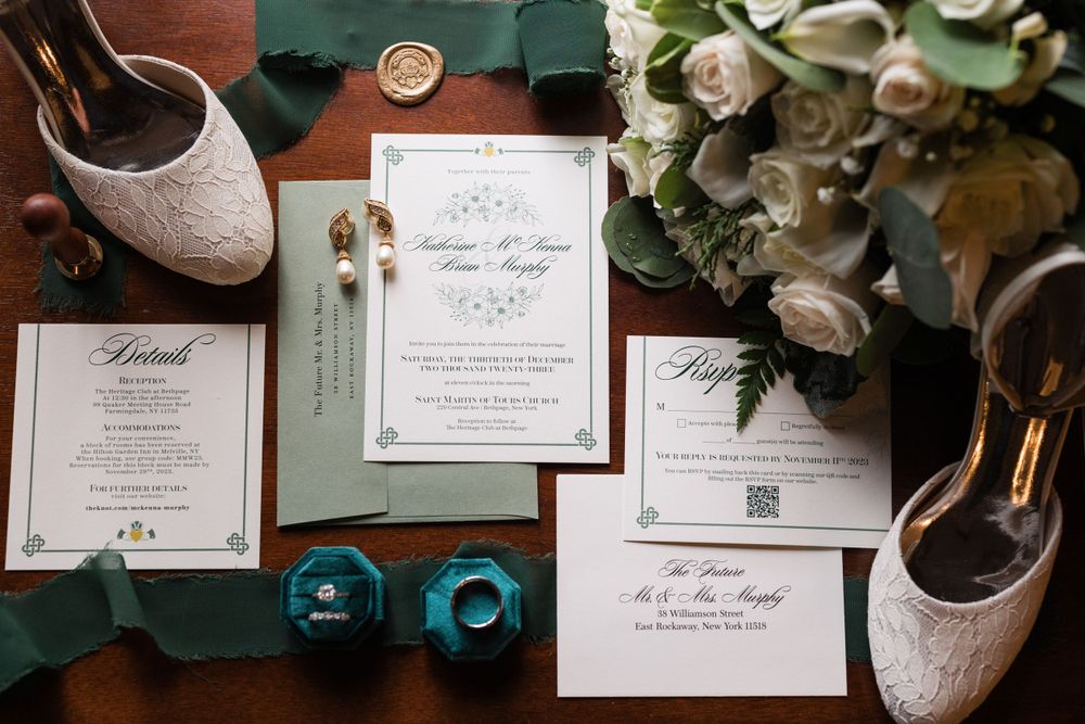 Wedding stationery and sparkly bridal shoes arranged with white roses on a dark green surface.