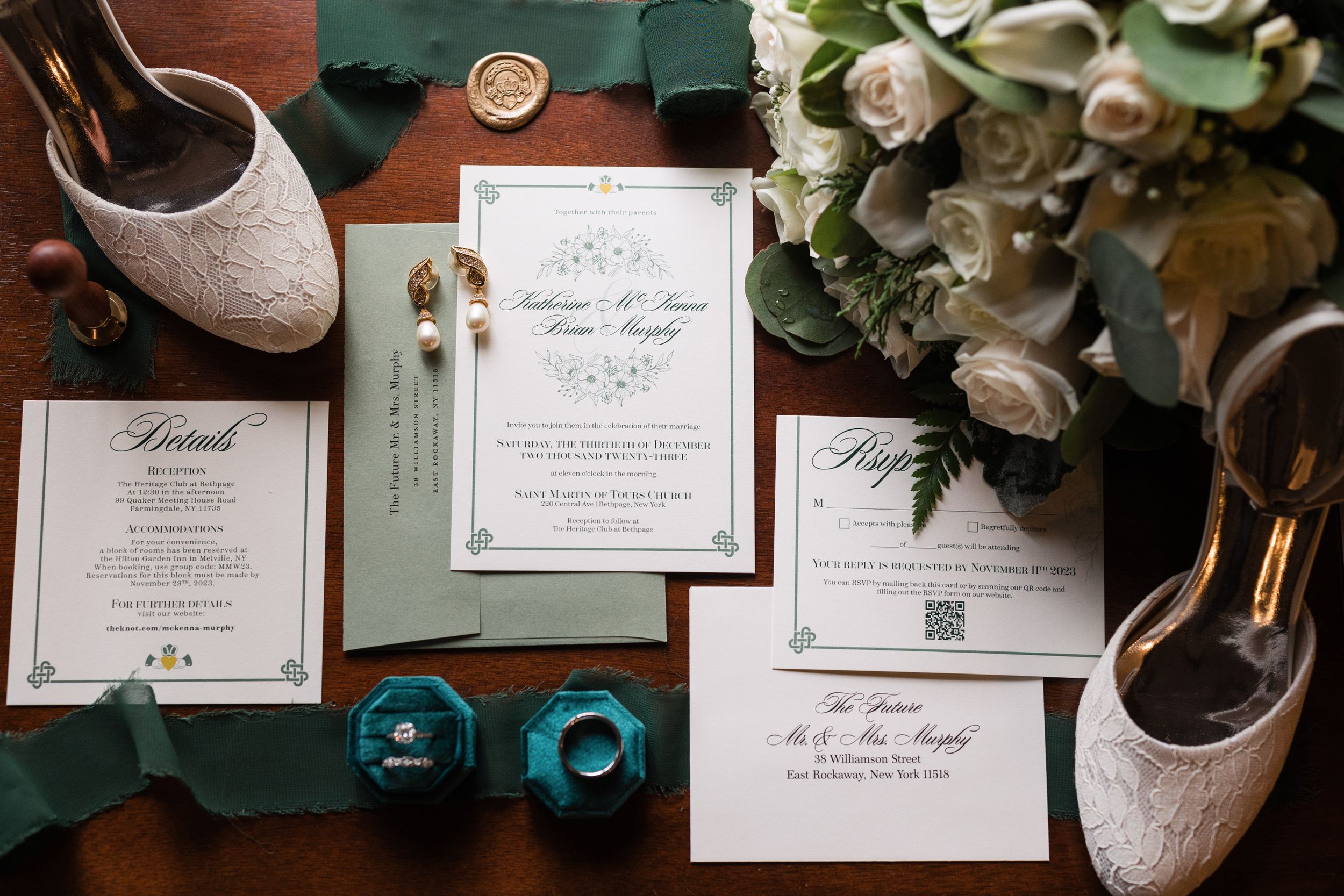 Wedding stationery and sparkly bridal shoes arranged with white roses on a dark green surface.
