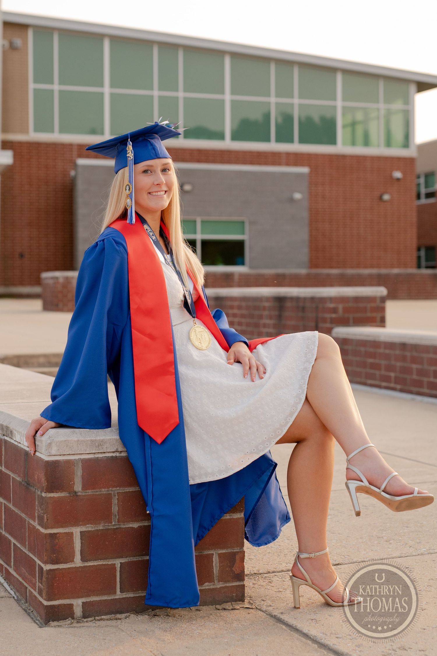 Graduate in blue cap and gown with red tie sits on brick steps outside modern school building for graduation photos.