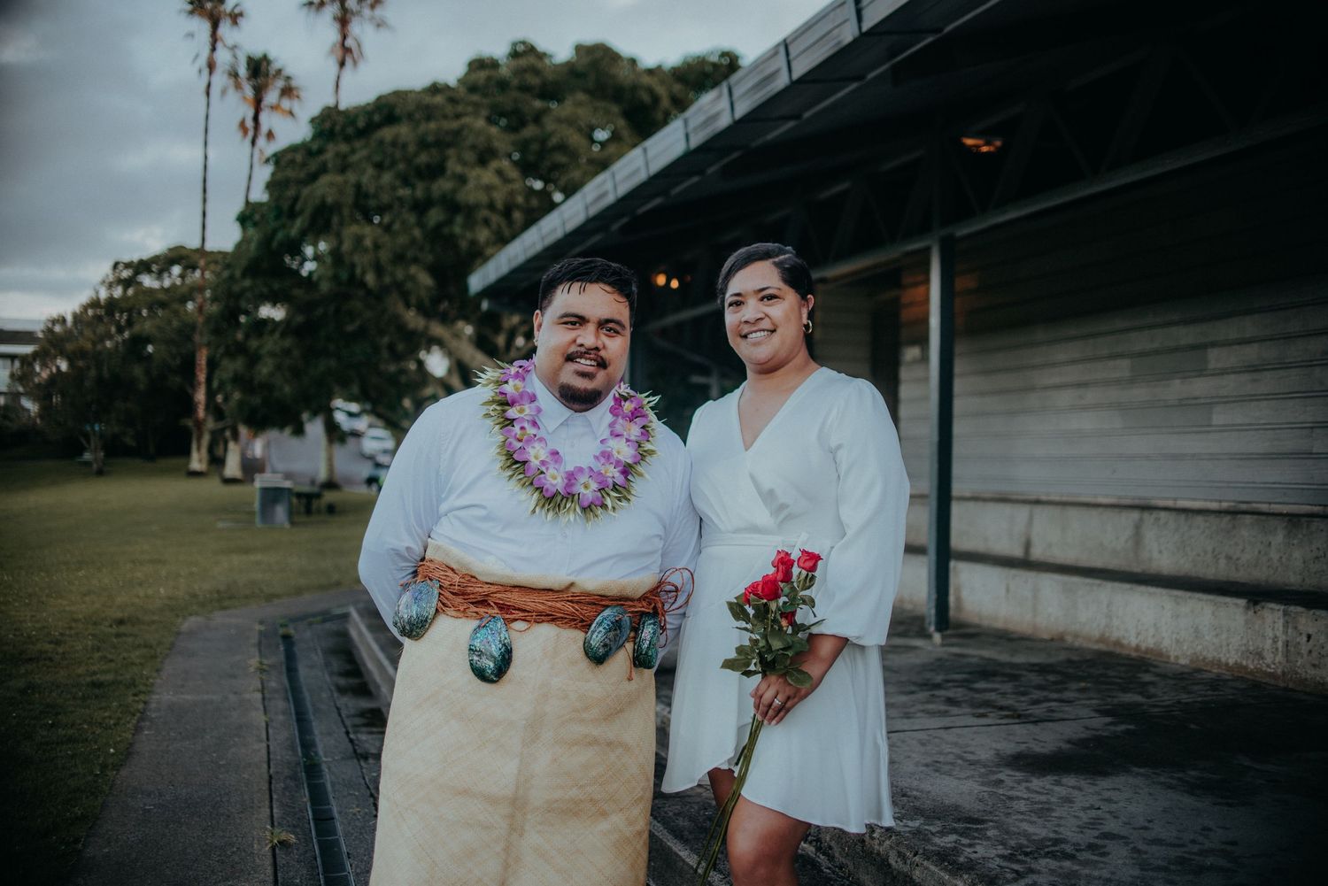 Couple in traditional Polynesian wedding attire pose together outdoors holding red flowers.