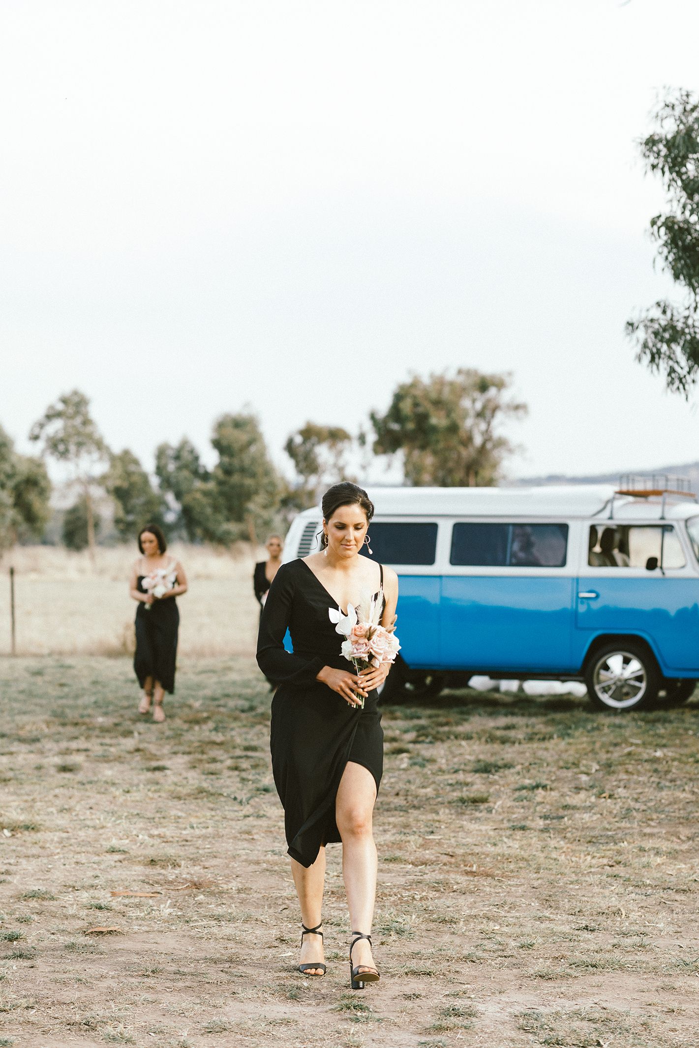 Bridesmaids walking across field with vintage blue Volkswagen van in background.