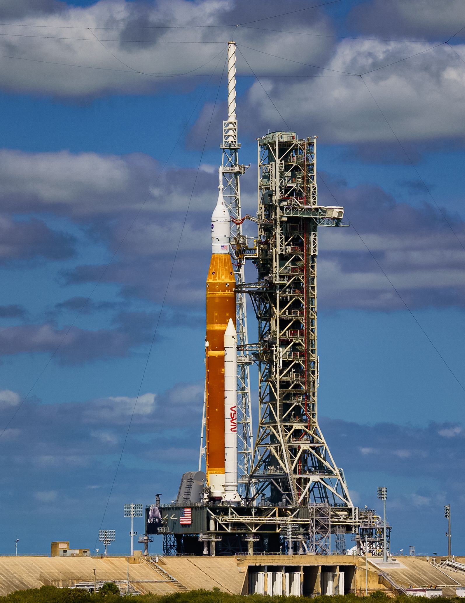 NASA's orange and white SLS rocket stands tall on the launch pad against a cloudy blue sky.
