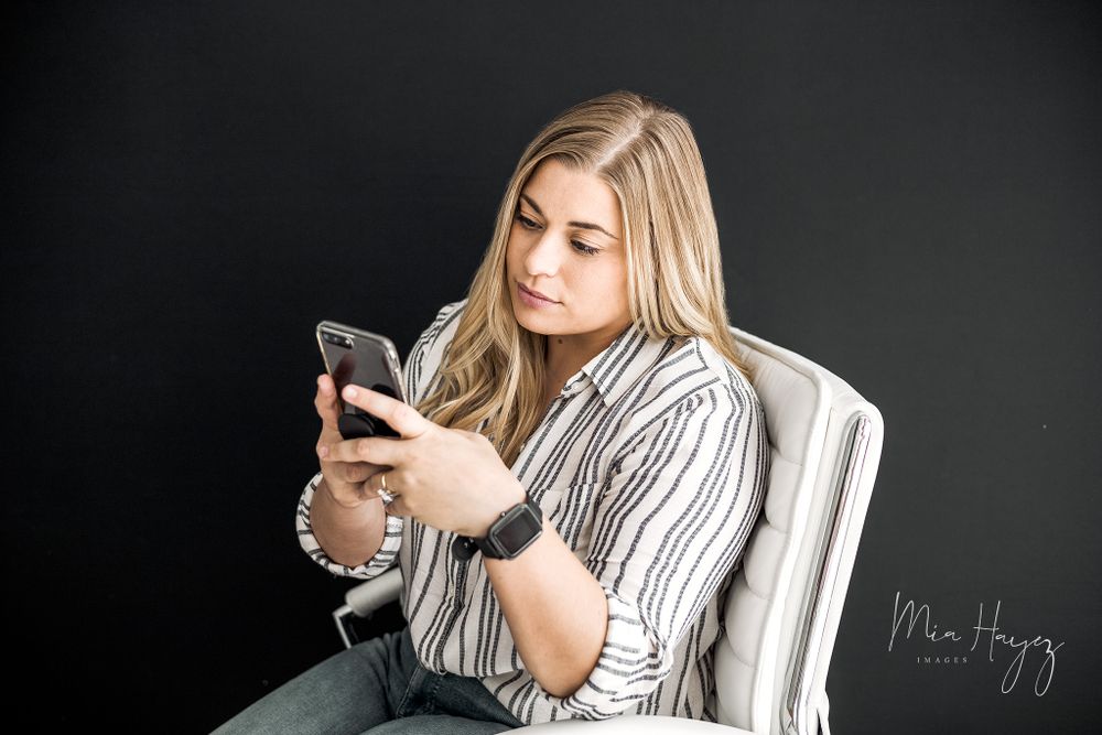 Person in striped top using mobile phone while seated in white chair against dark background.